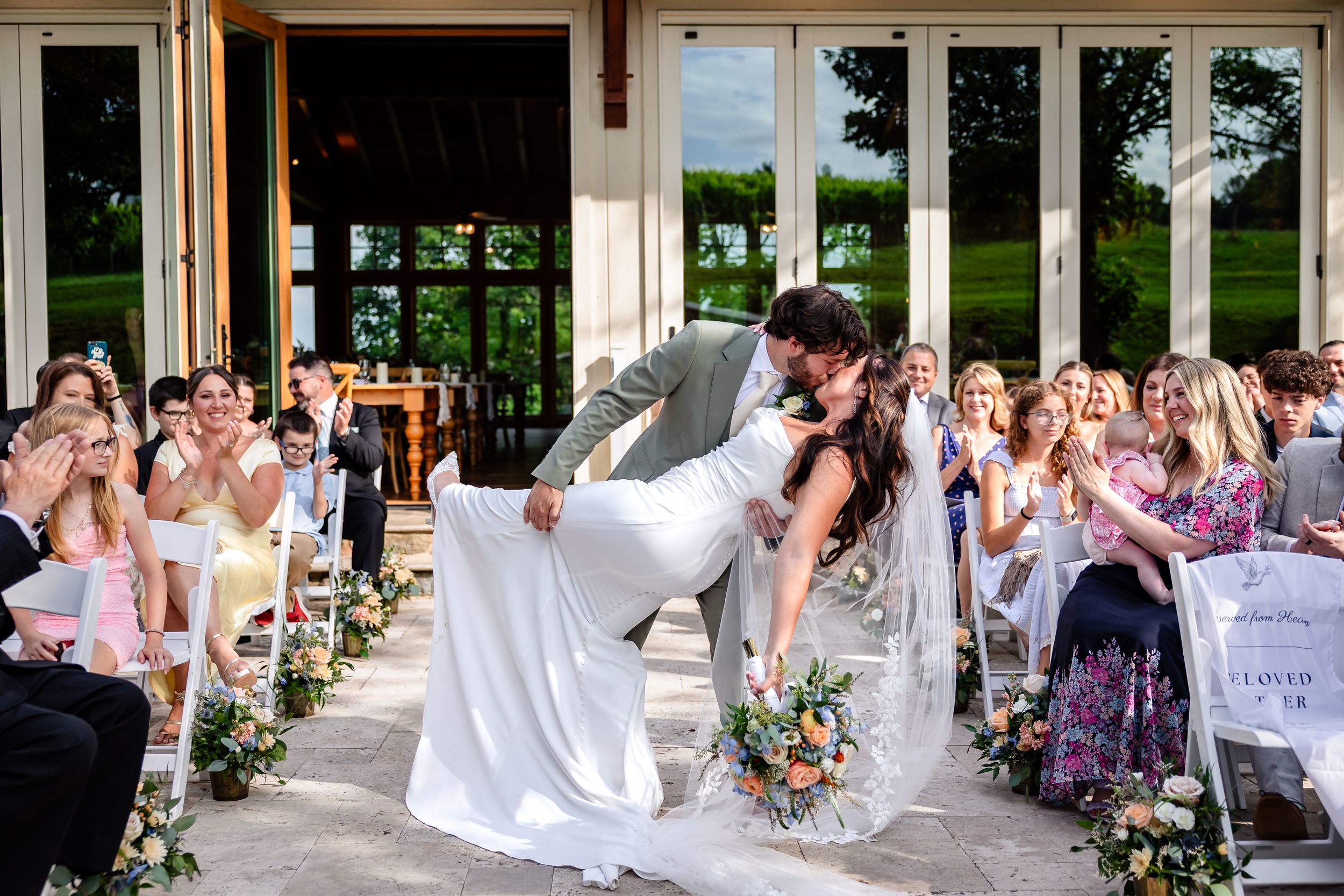 Groom dipping bride for kiss while guests cheer along decorated outdoor wedding aisle.