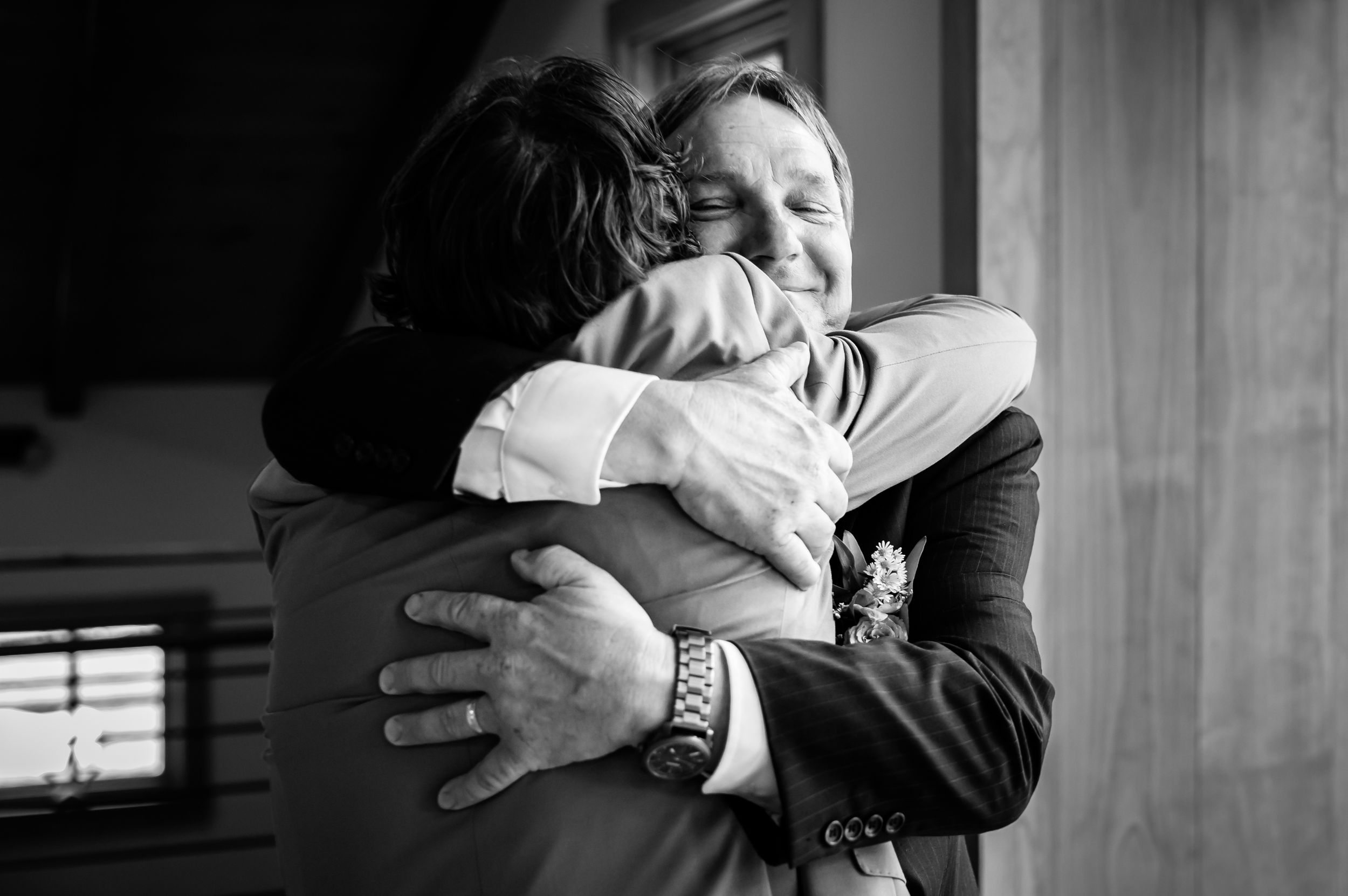 Emotional black and white photo of groom sharing a heartfelt hug with a family member before wedding.