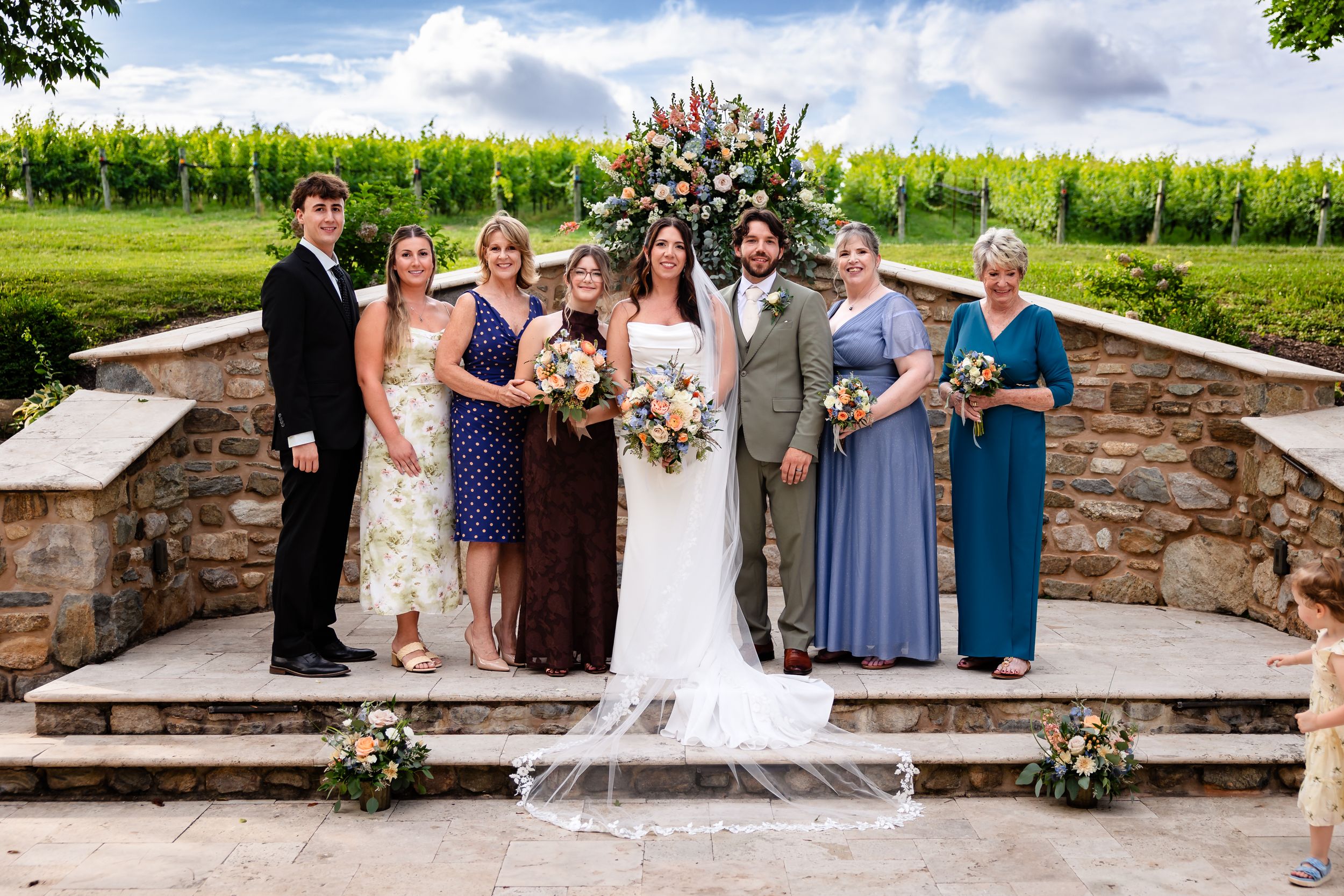 Wedding party poses at stone ceremony altar surrounded by vineyard with floral arch backdrop.