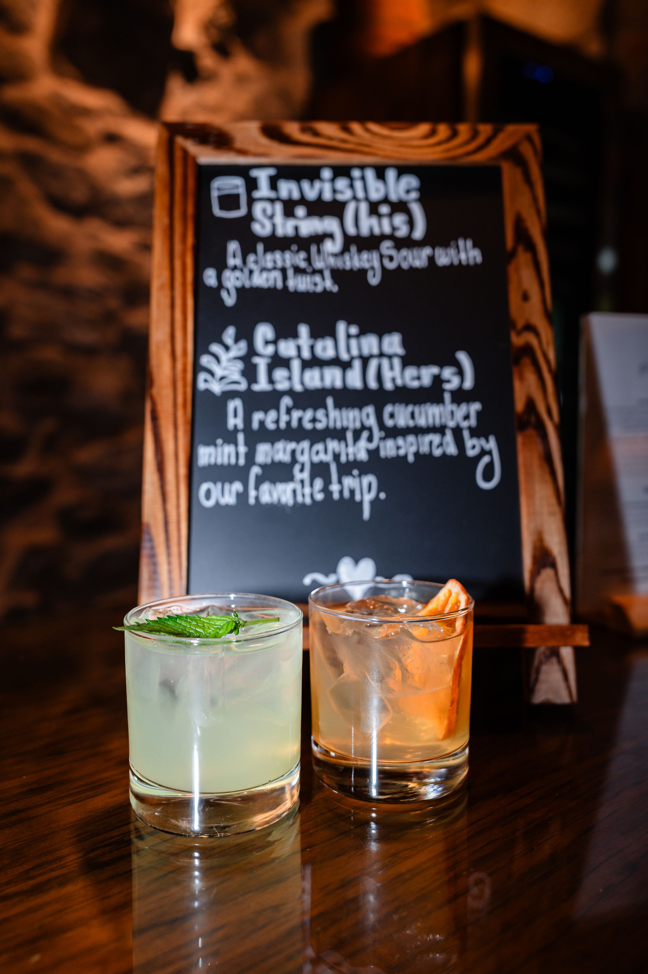 Two signature wedding cocktails on a wooden bar beside a chalkboard menu featuring Invisible String and Catalina Island drinks.
