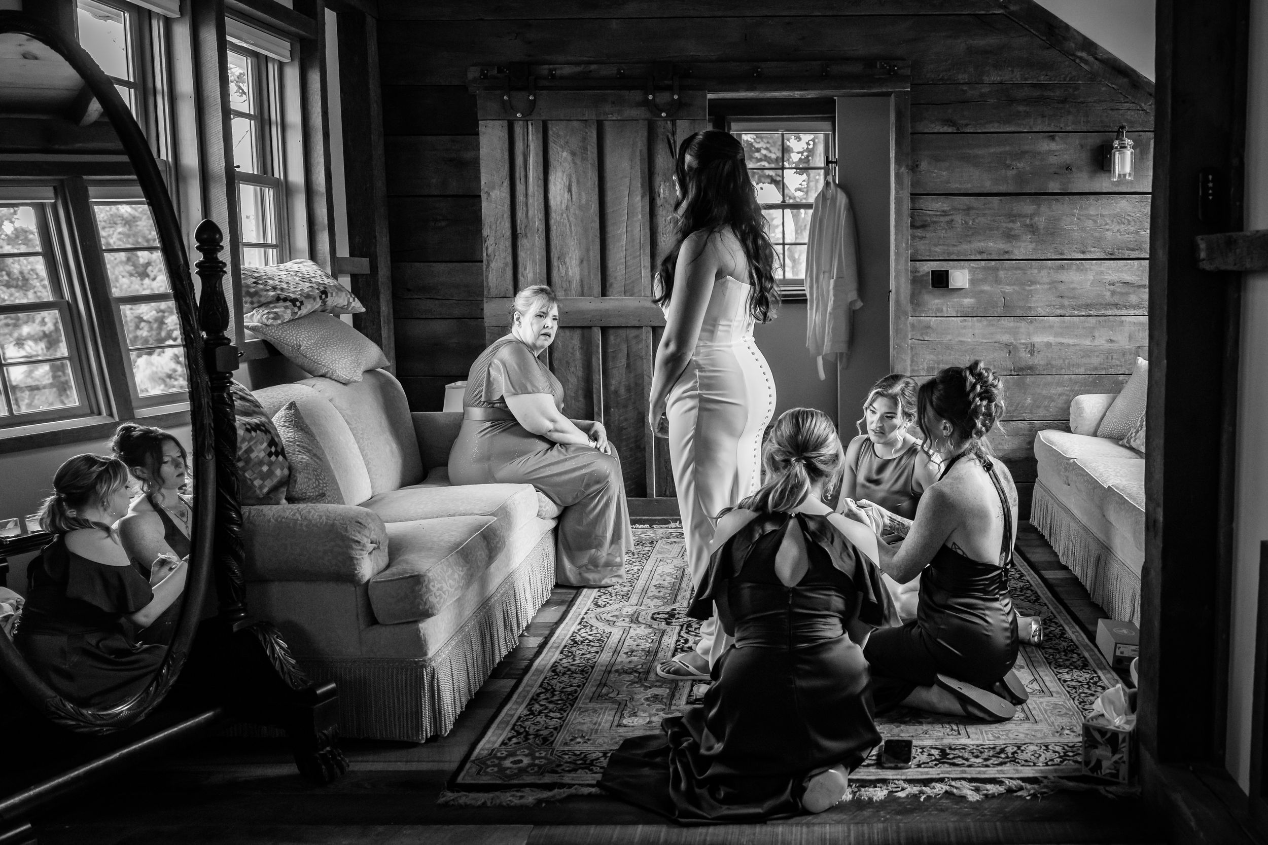 Black and white photo of bride in wedding dress surrounded by bridesmaids getting ready in a rustic wood-paneled room.