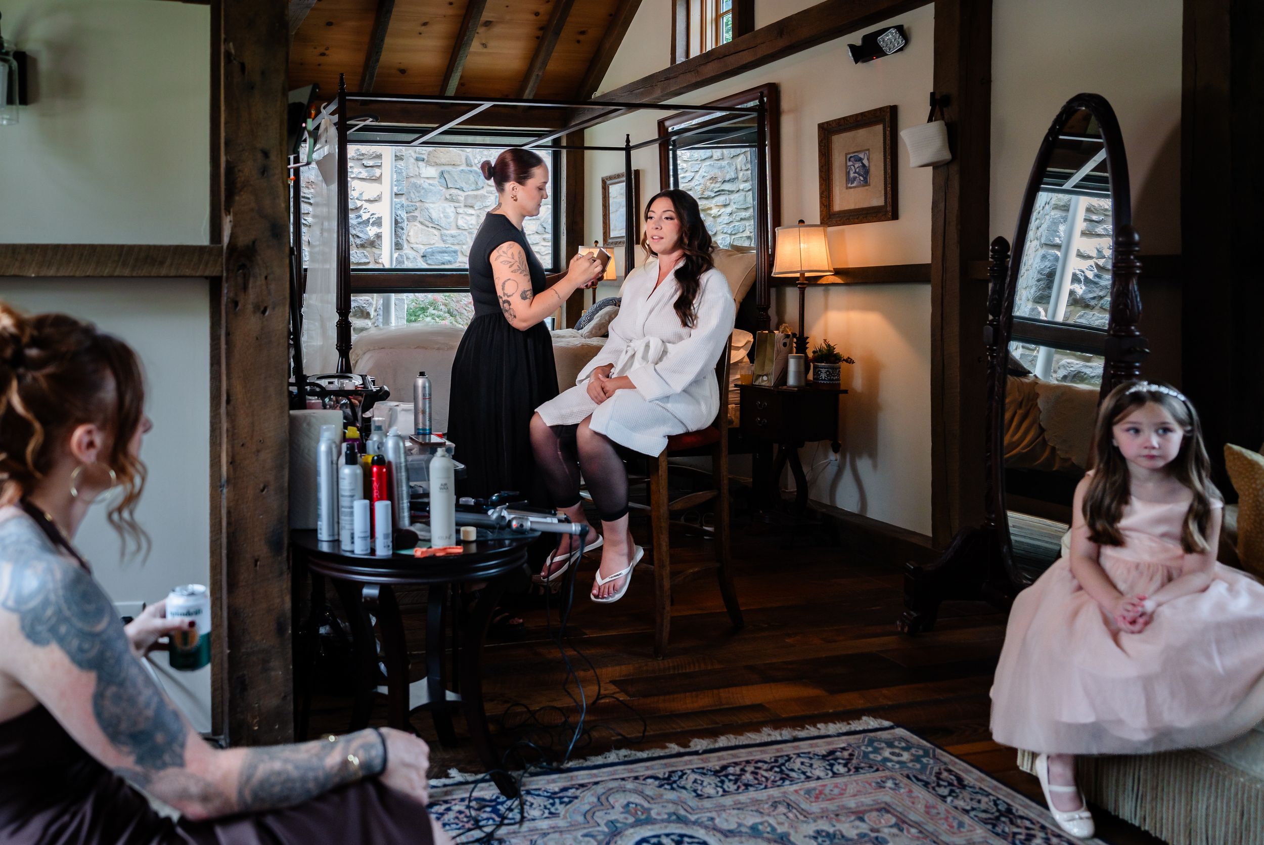 Bride getting makeup done in a rustic cabin while a flower girl in pink watches nearby.