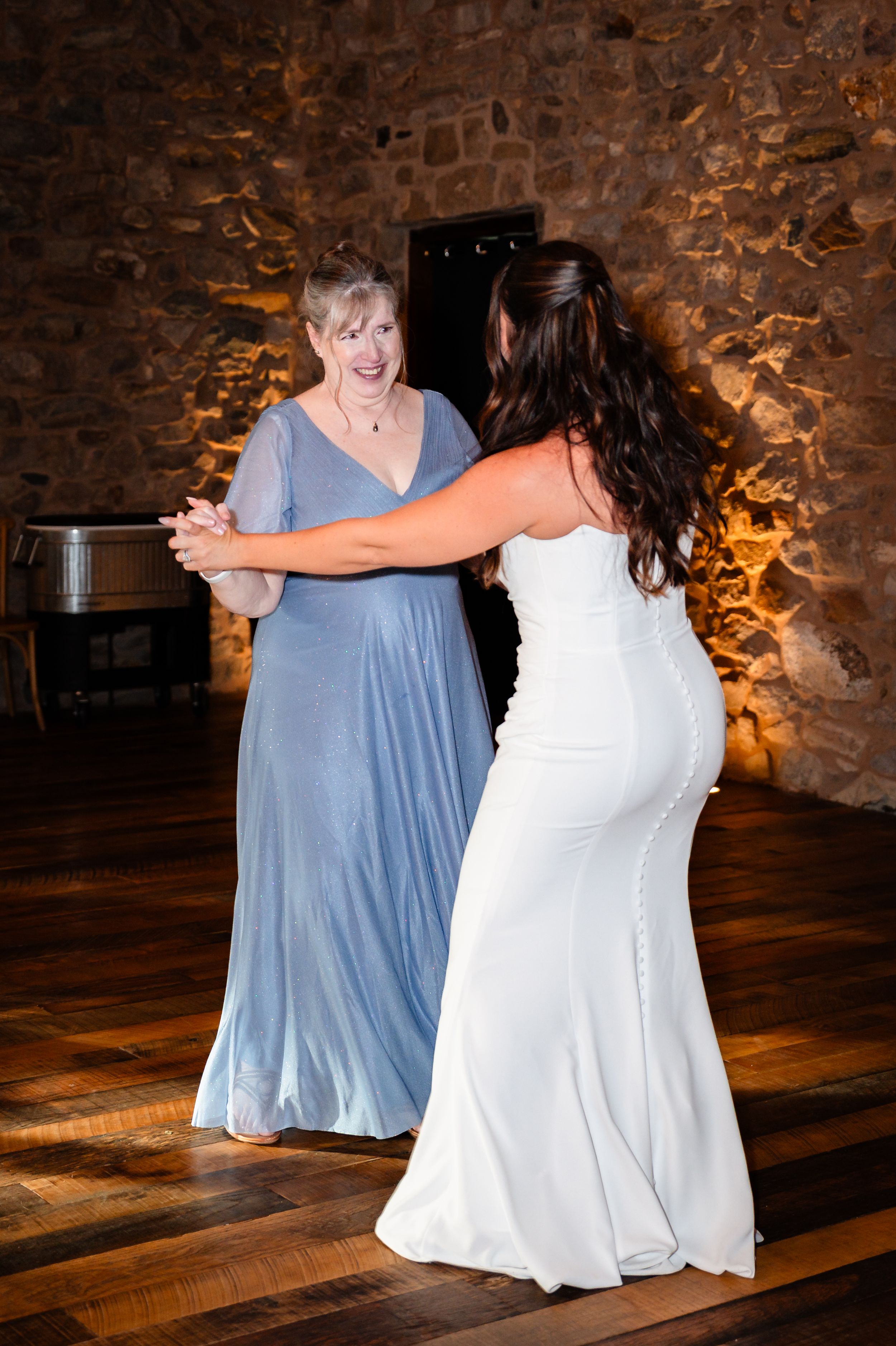 Bride in white gown dancing with older woman in blue dress at rustic stone wall wedding reception.
