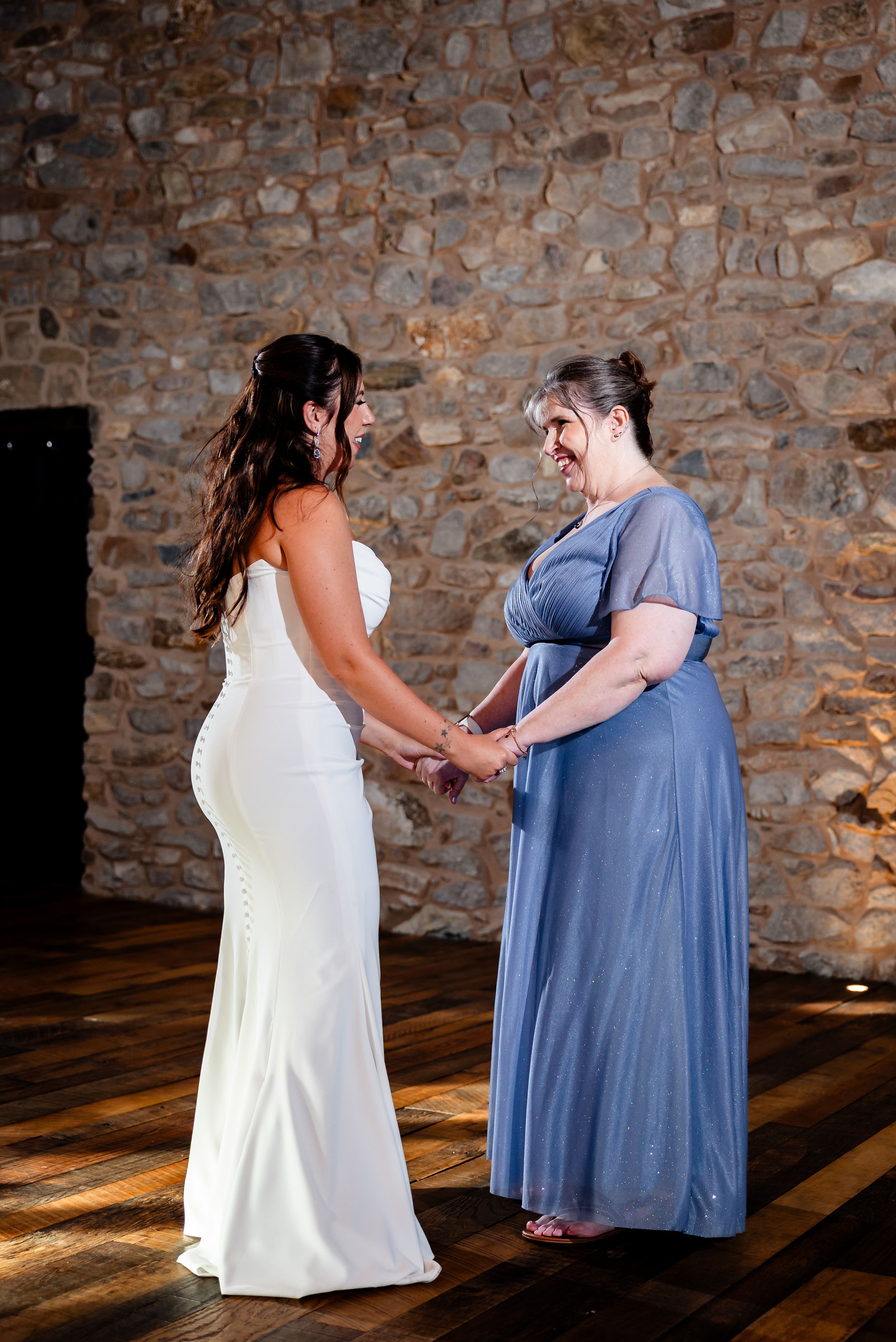 Bride in white gown holds hands with woman in blue dress, sharing a tender moment on a wooden dance floor.
