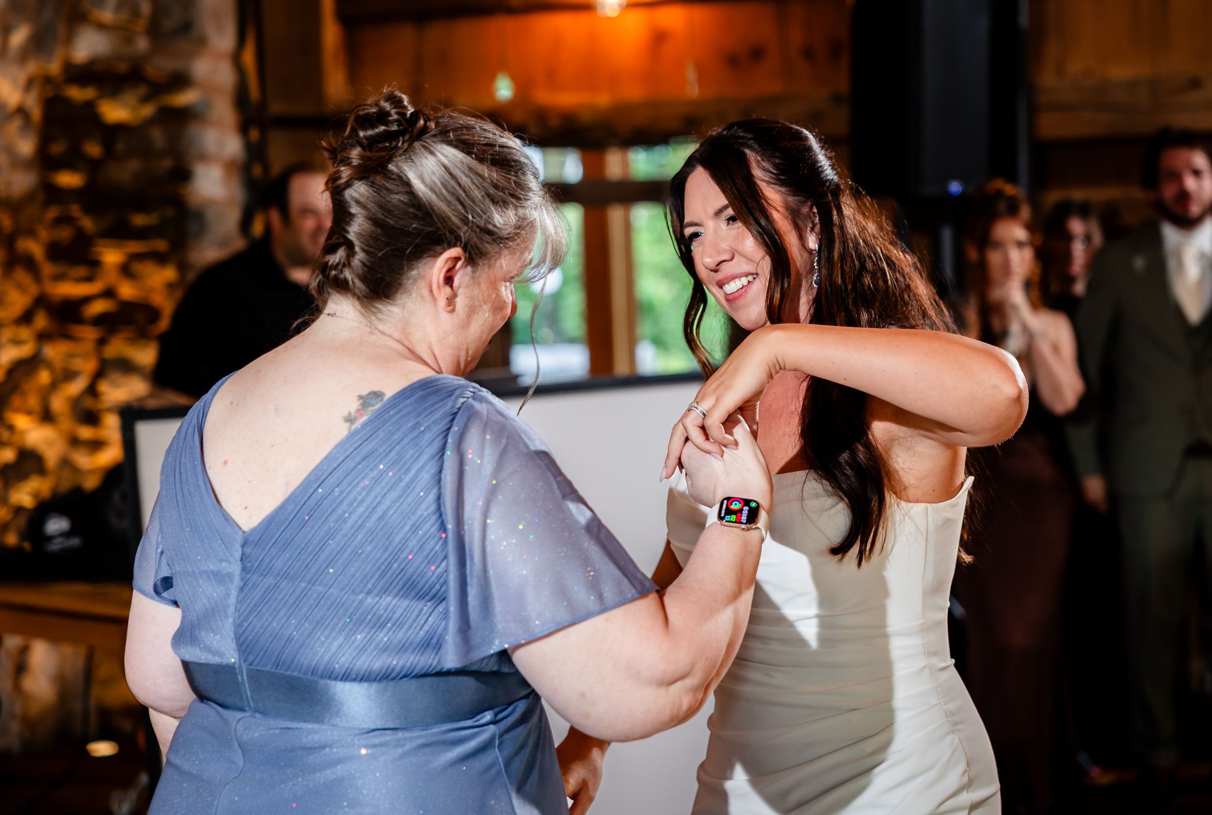 Bride and older woman share a joyful dance together at a rustic wedding reception venue.