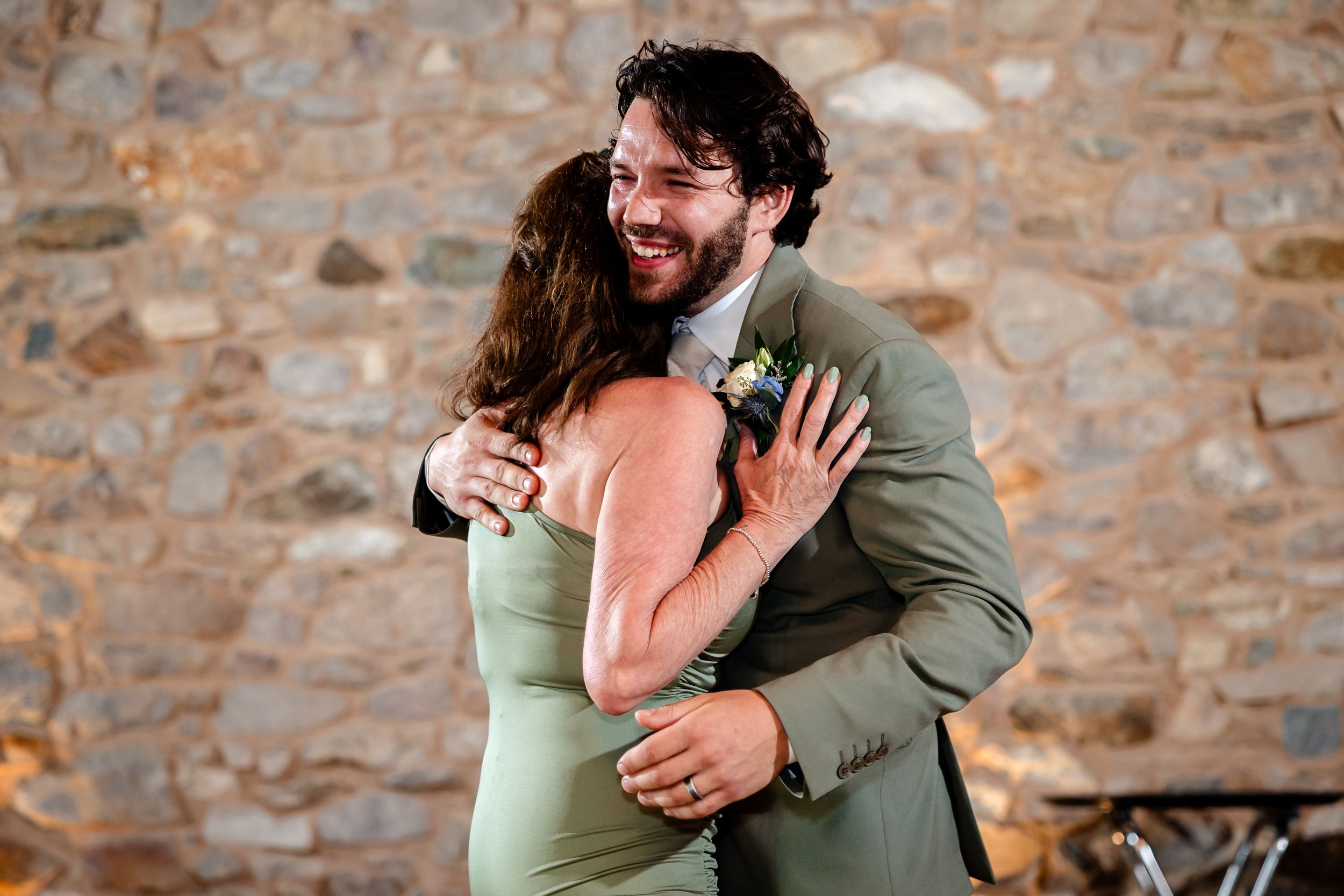 Joyful couple sharing a warm embrace during wedding reception dance in front of rustic stone wall.