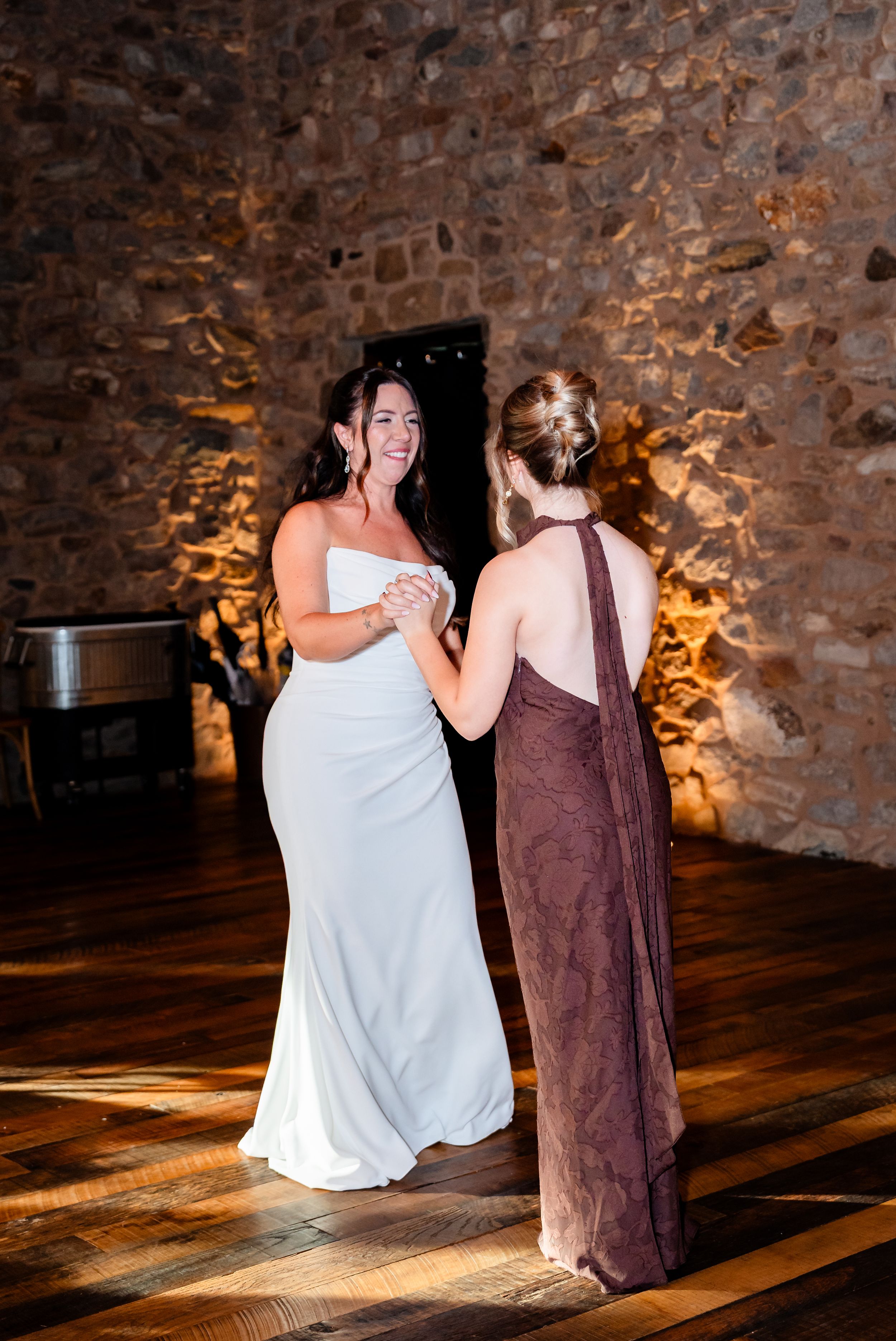 Bride in white gown dancing with bridesmaid in mauve dress in rustic stone venue.