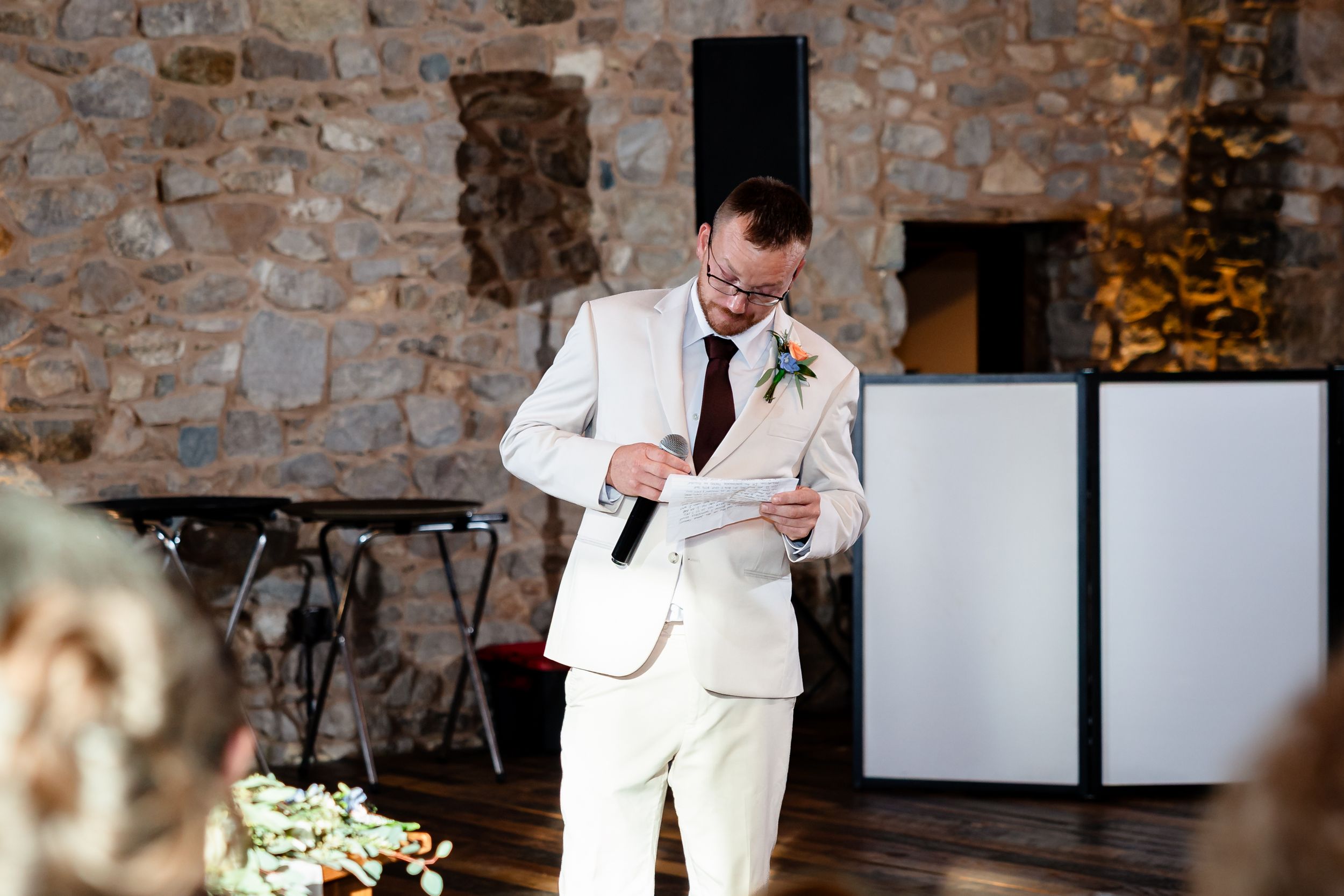 Groom in white suit reading vows from paper while holding microphone at rustic stone wall wedding venue.