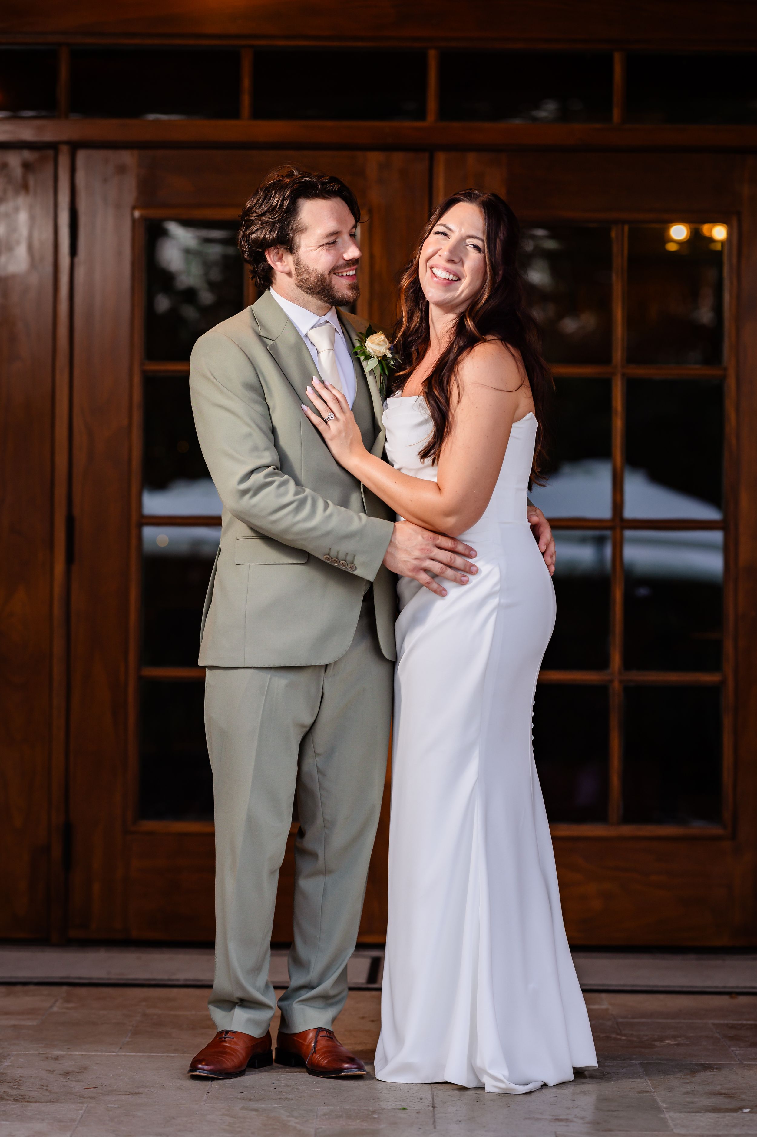 Laughing bride in white gown embraced by groom in sage suit before wooden doors.