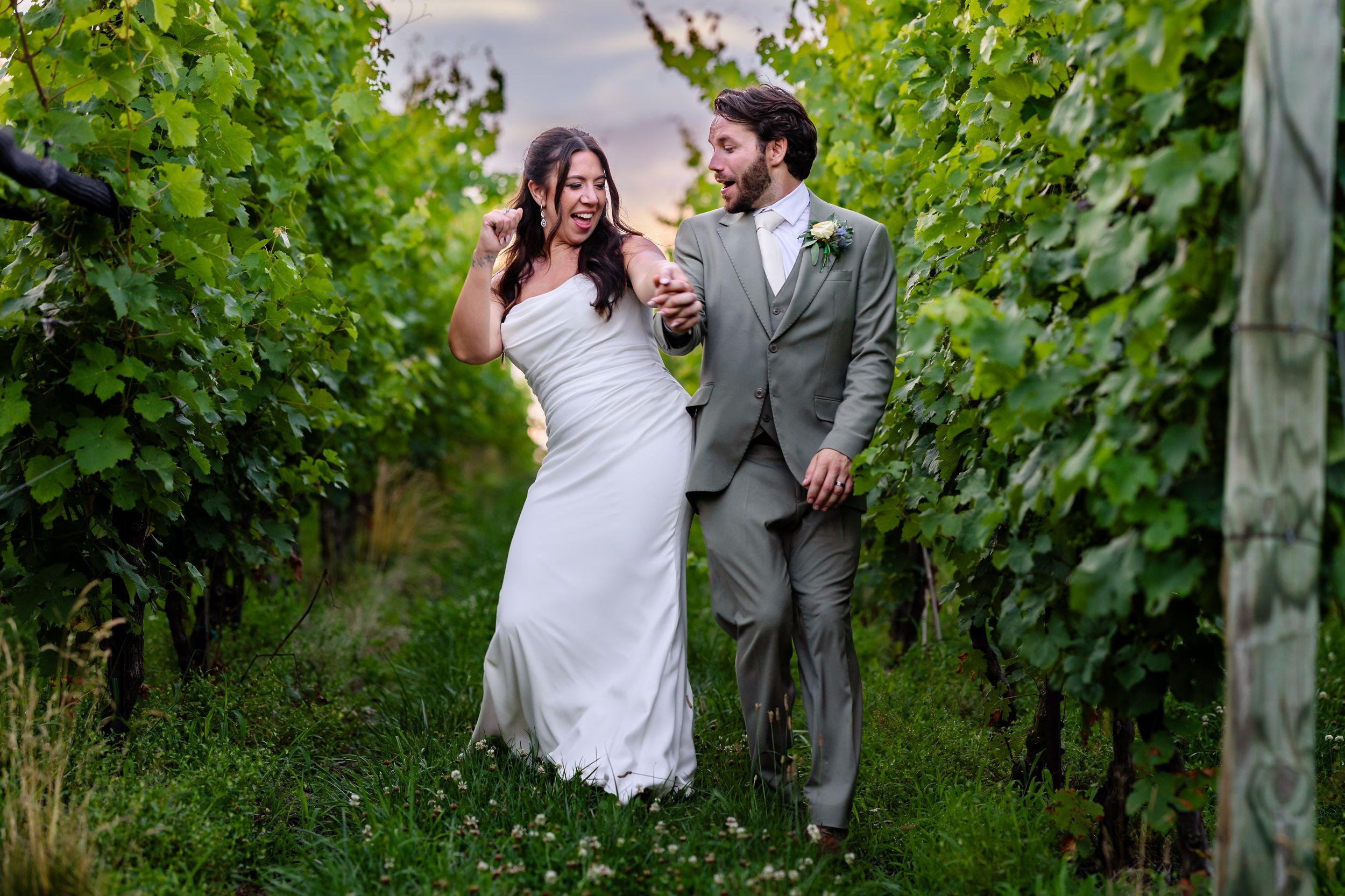 Bride and groom walk through vineyard rows holding champagne glasses at sunset wedding.