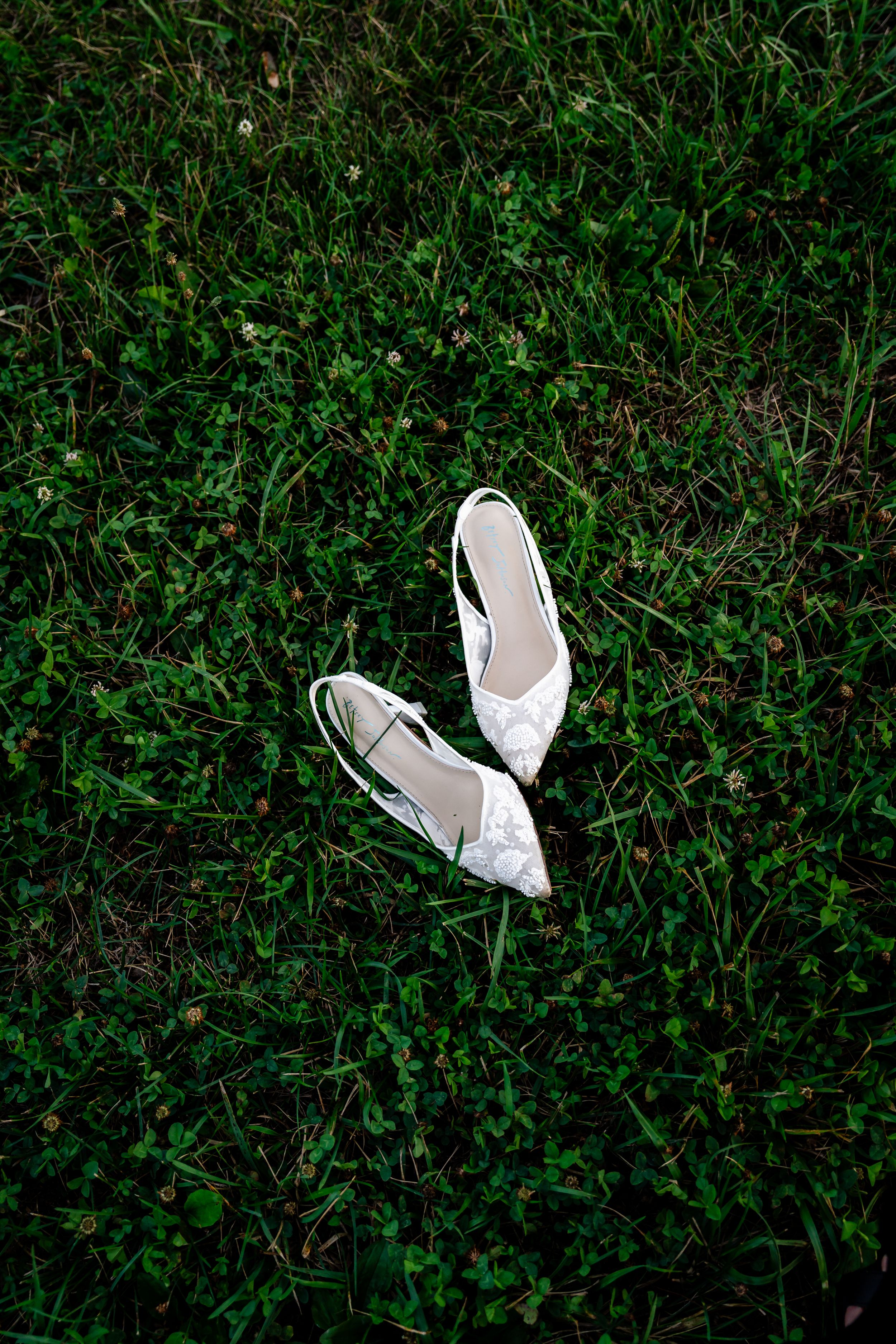 White lace bridal heels laid on lush green grass for wedding day detail shot.