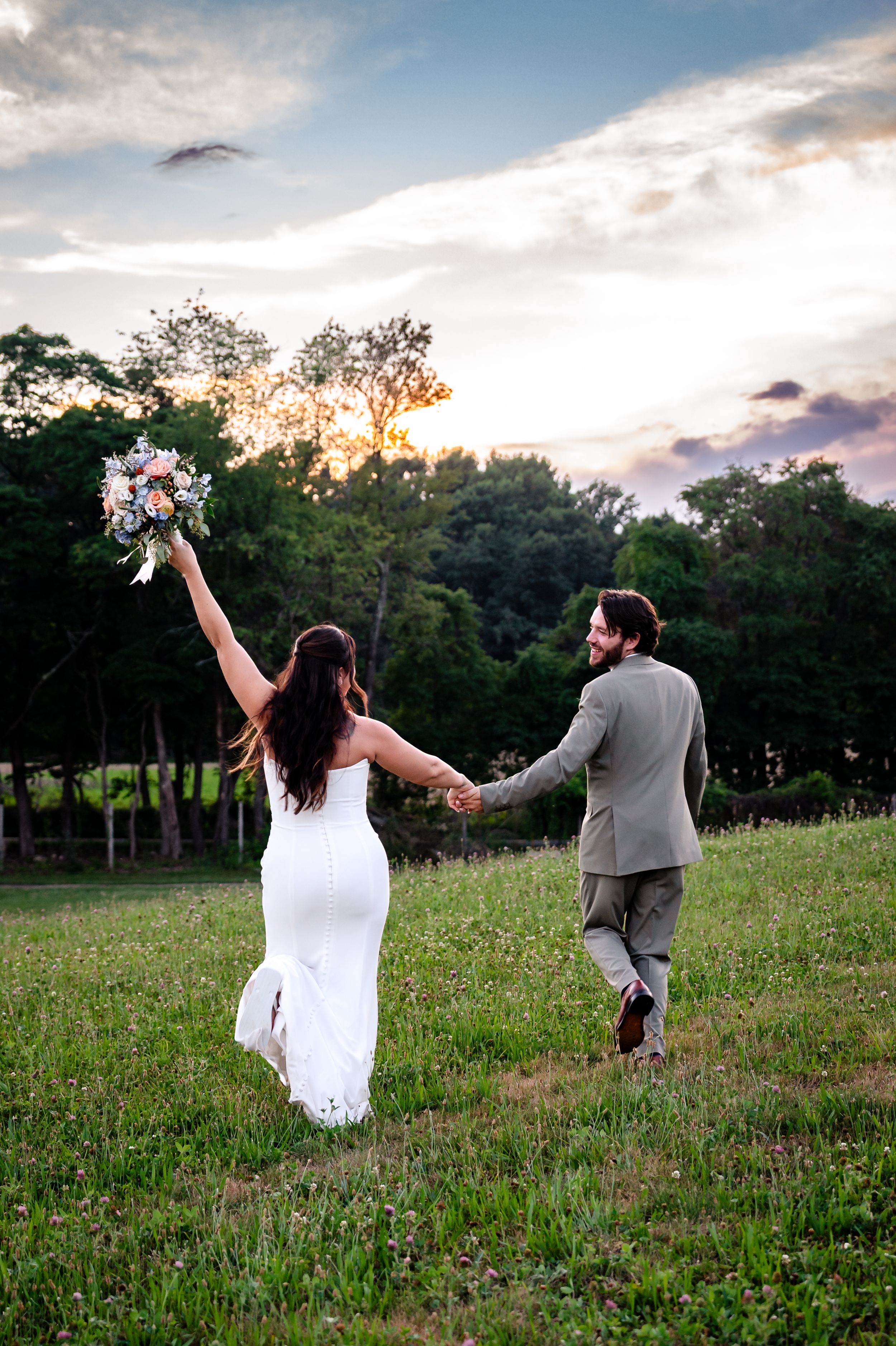 Bride raises bouquet while holding hands with groom in gray suit, walking through green field at golden sunset.