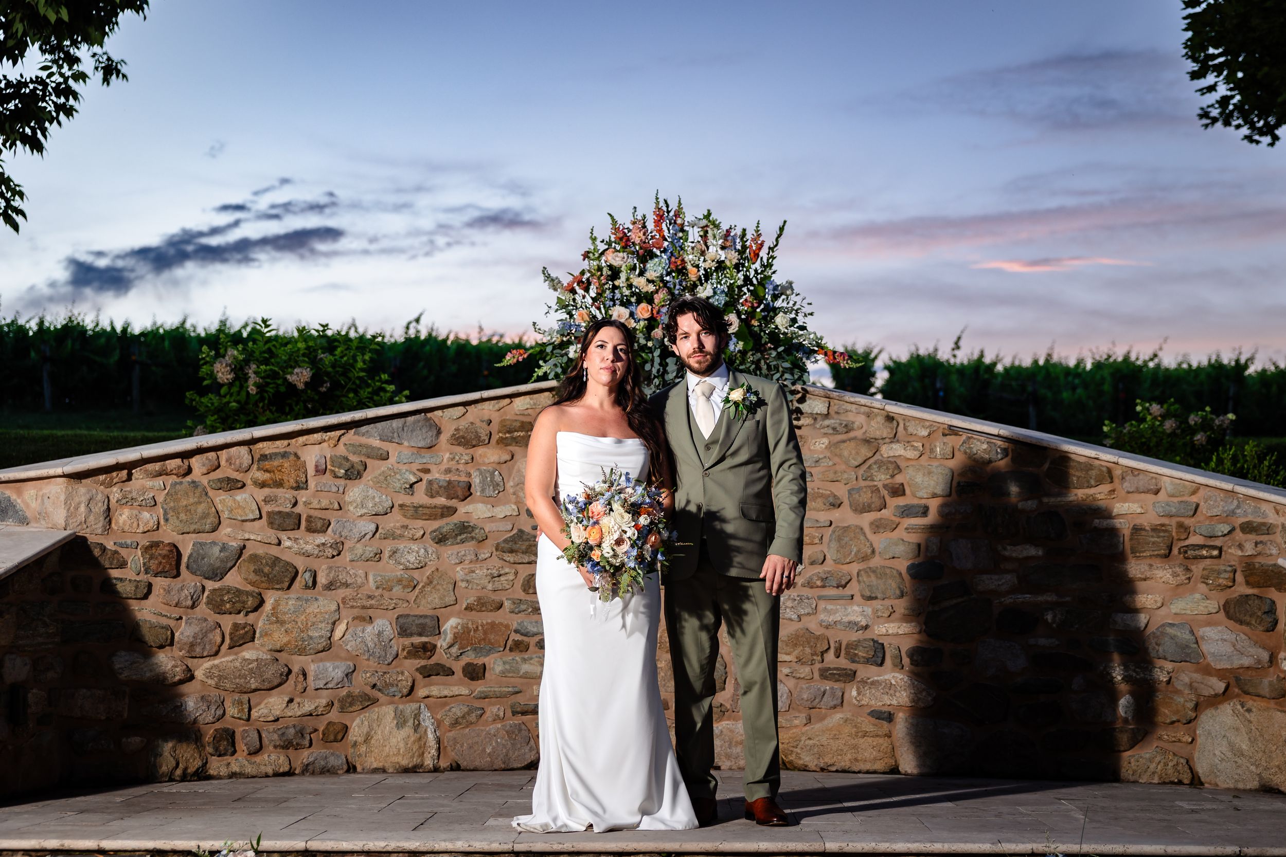 Bride and groom pose before a floral arch against a stone wall at sunset during their outdoor wedding ceremony.