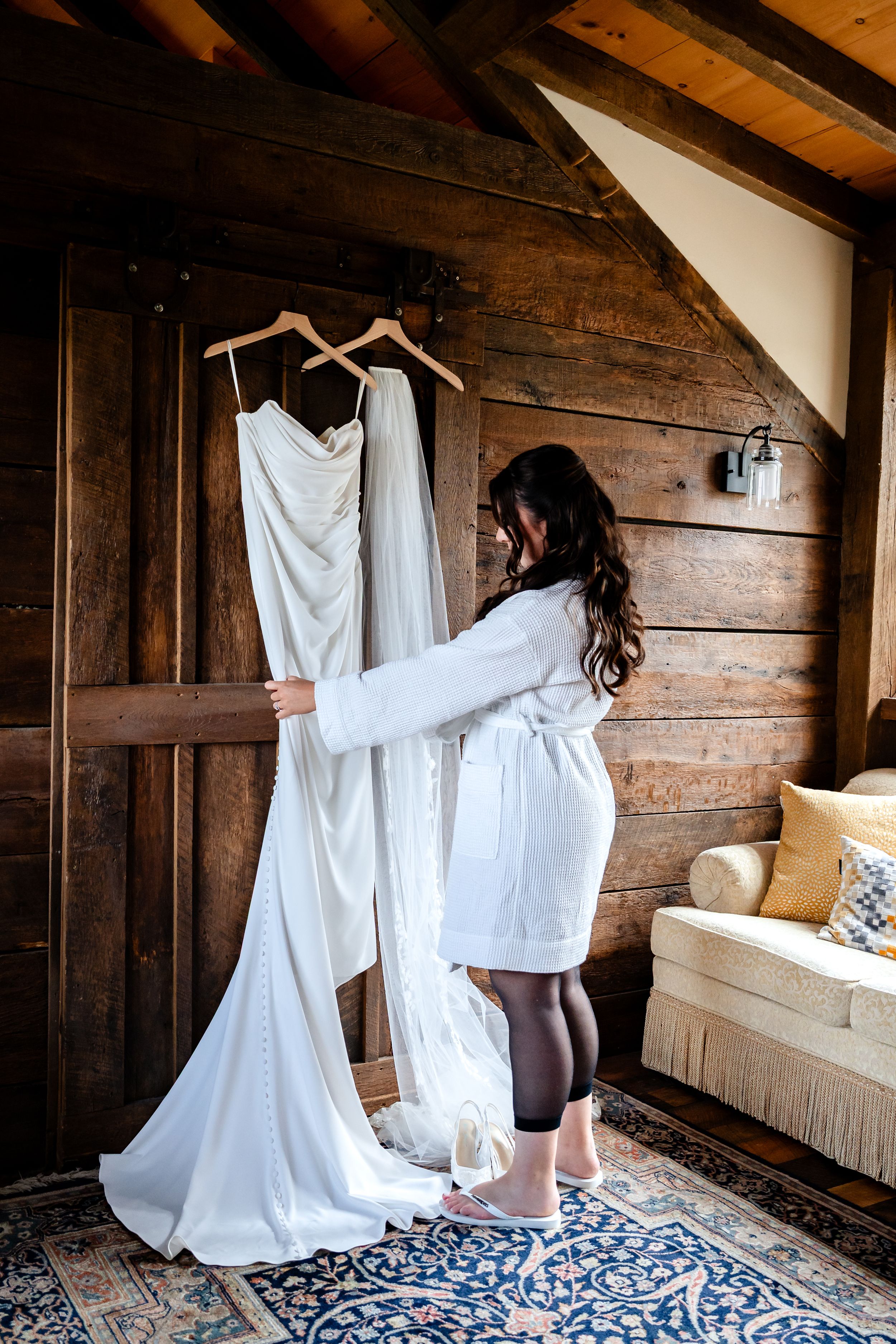 Bride in white robe admiring her wedding gown hanging on wooden hangers in a rustic barn venue.