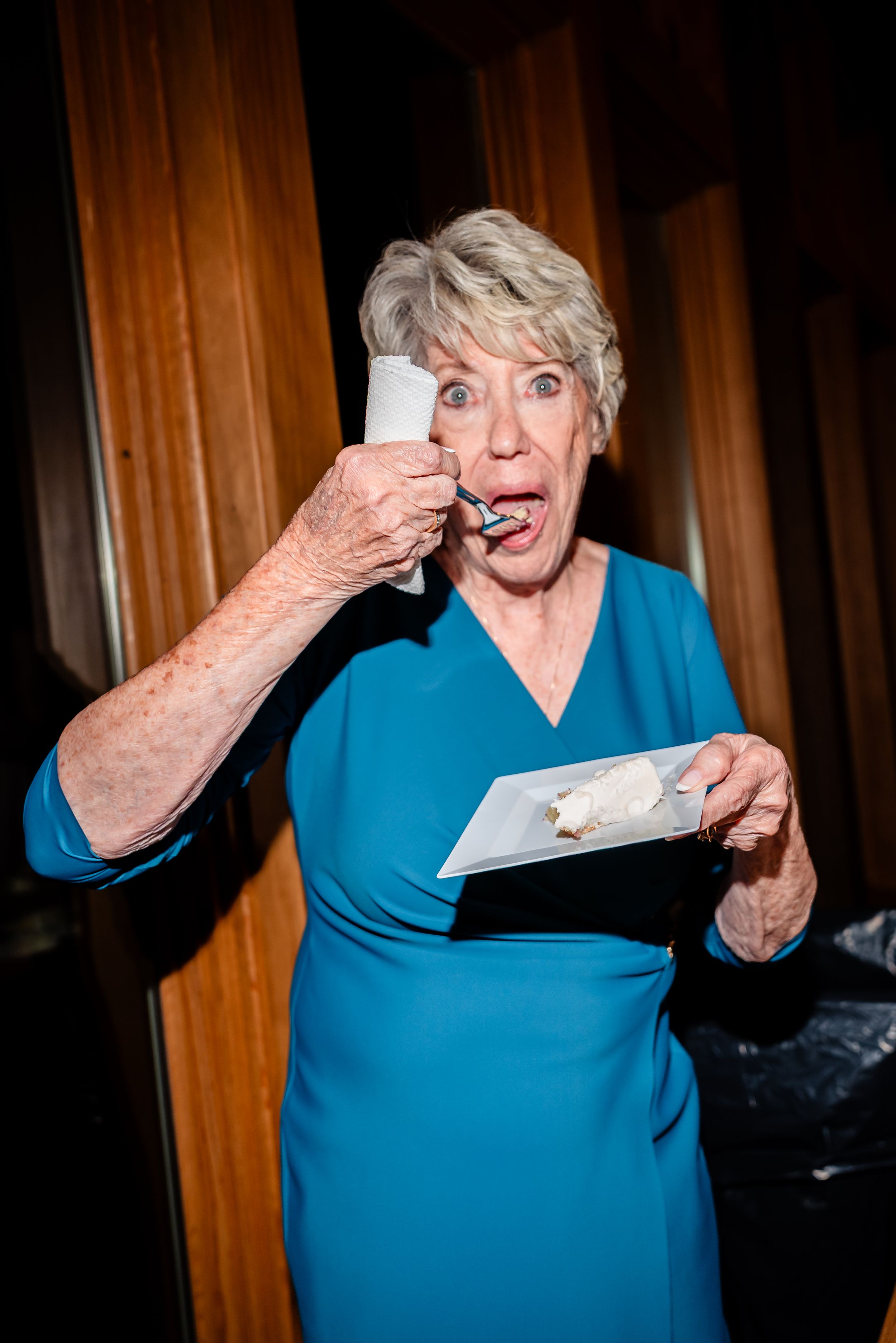 Elderly woman in blue dress enjoying wedding cake with surprised expression at reception.