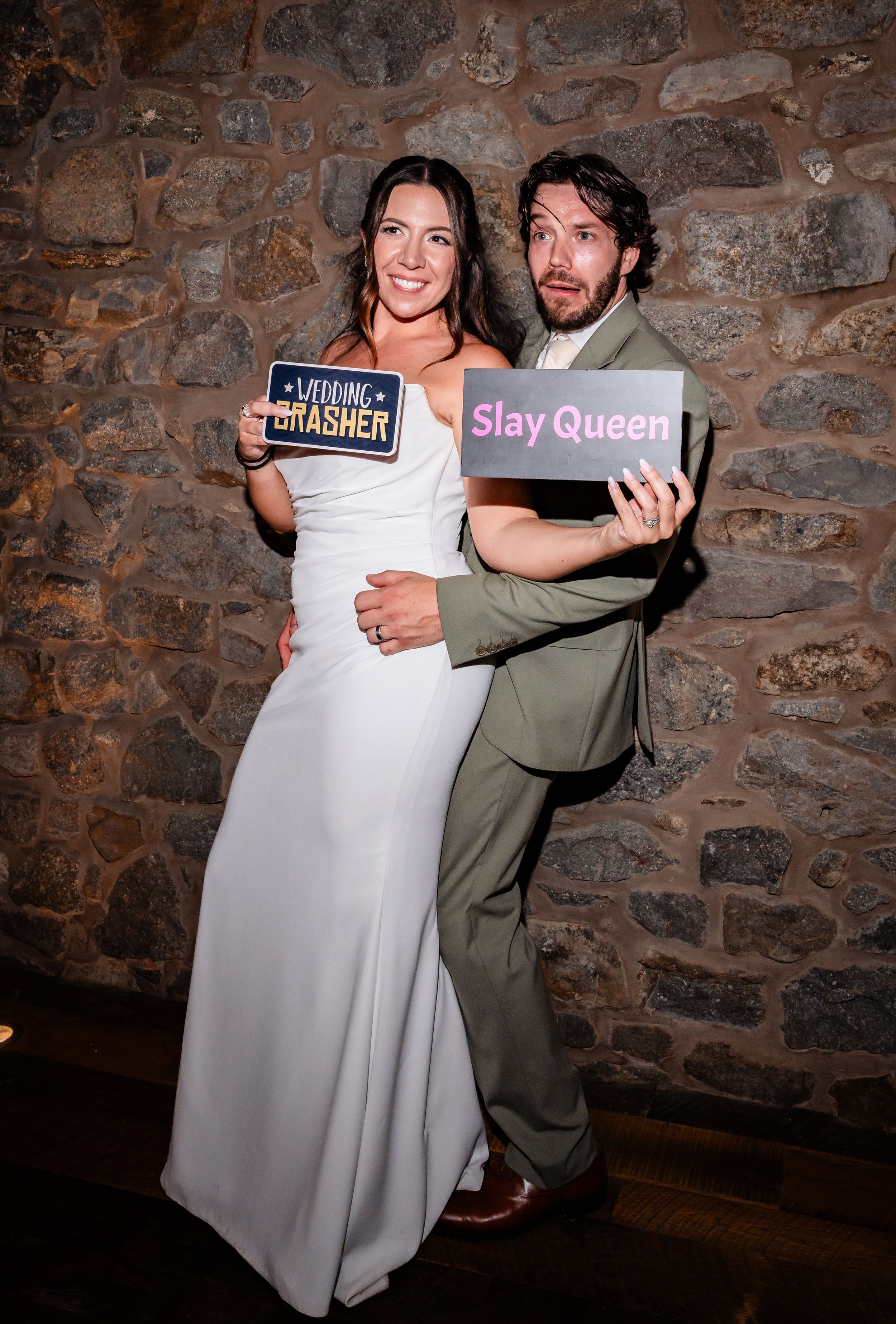 Bride and groom pose with fun photo booth signs at rustic stone wall wedding reception.