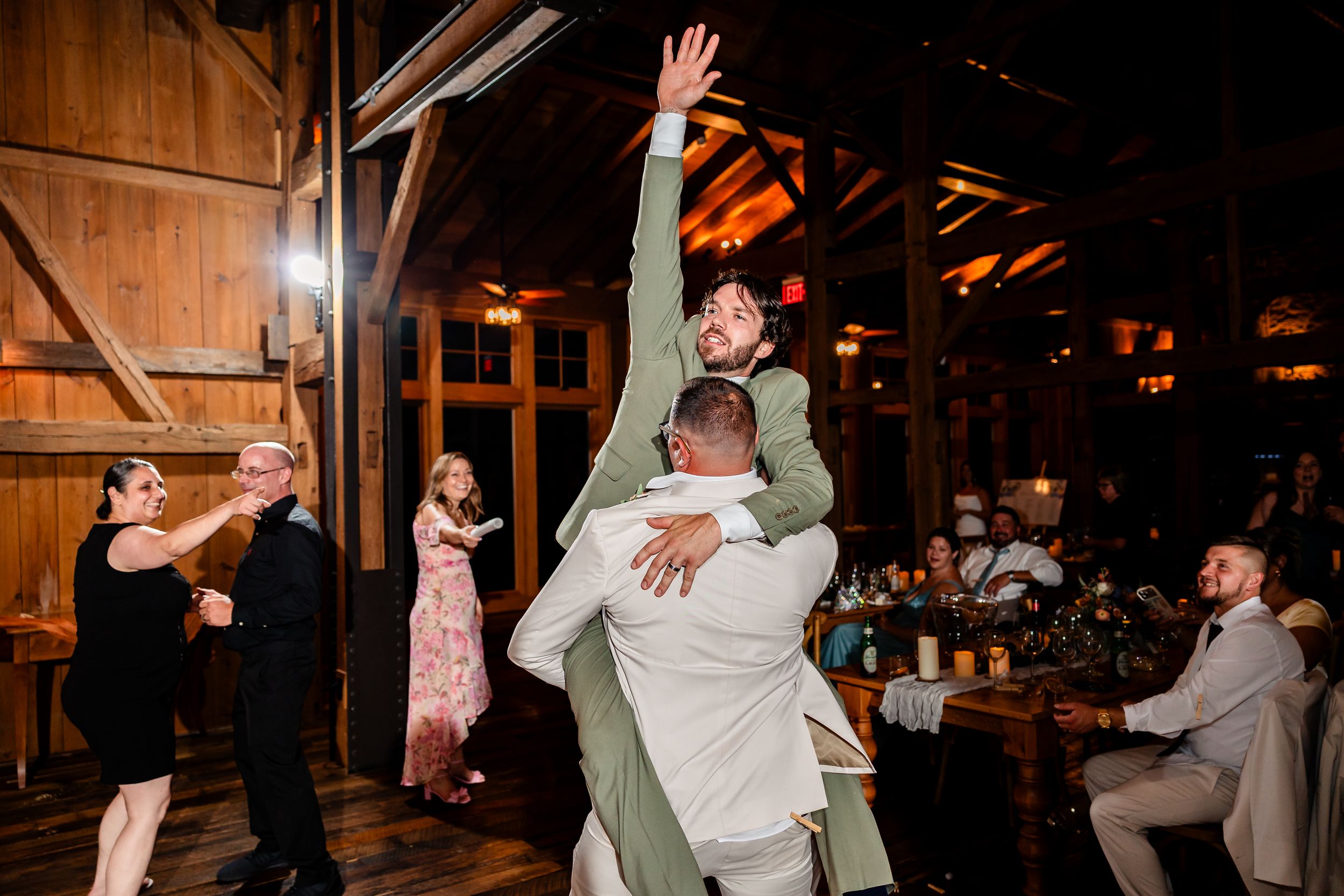 Groomsmen celebrate at rustic barn wedding reception with energetic dancing and lifting.