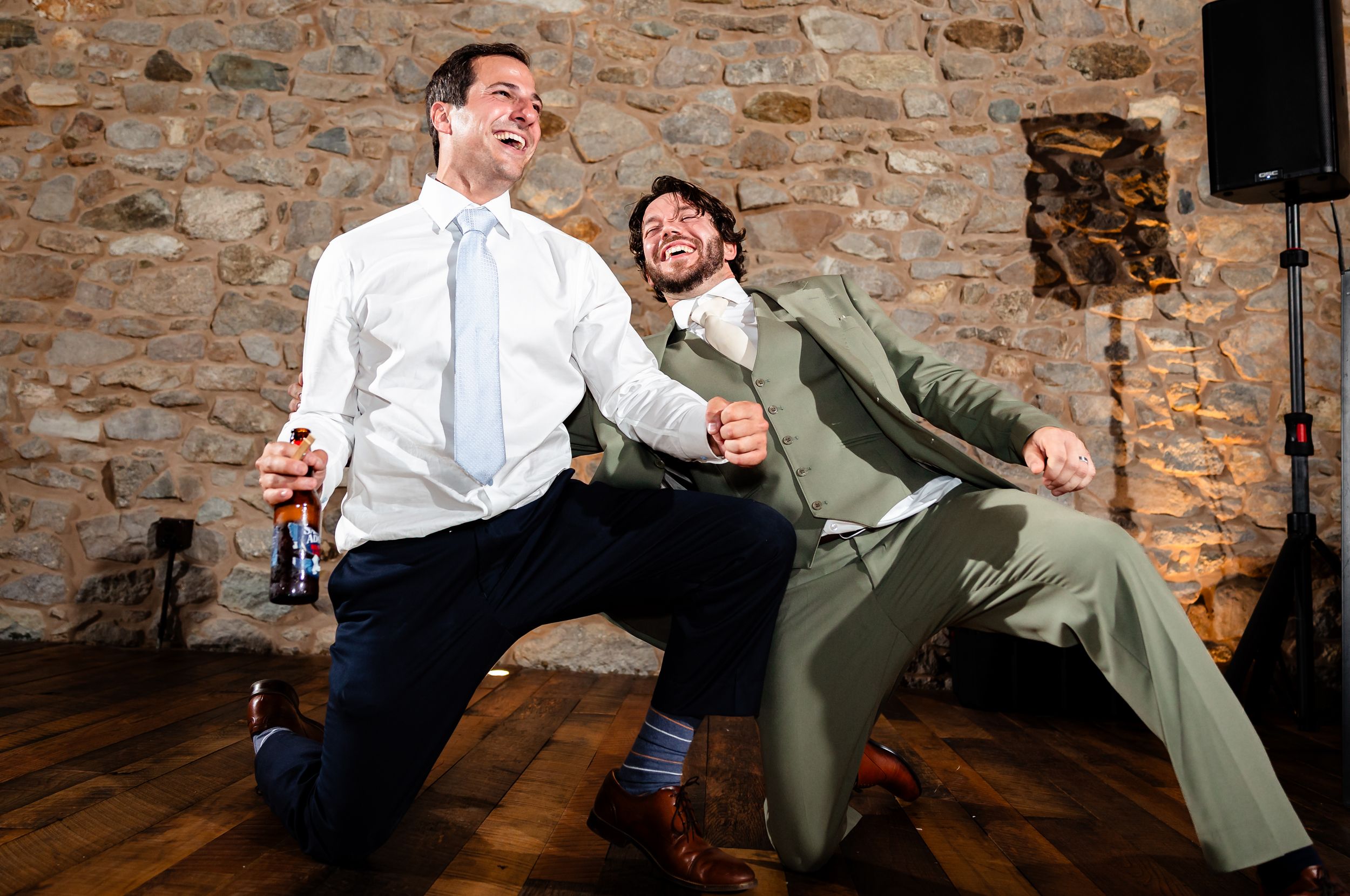 Two men dancing energetically at a wedding reception in a rustic stone-walled venue with hardwood floors.