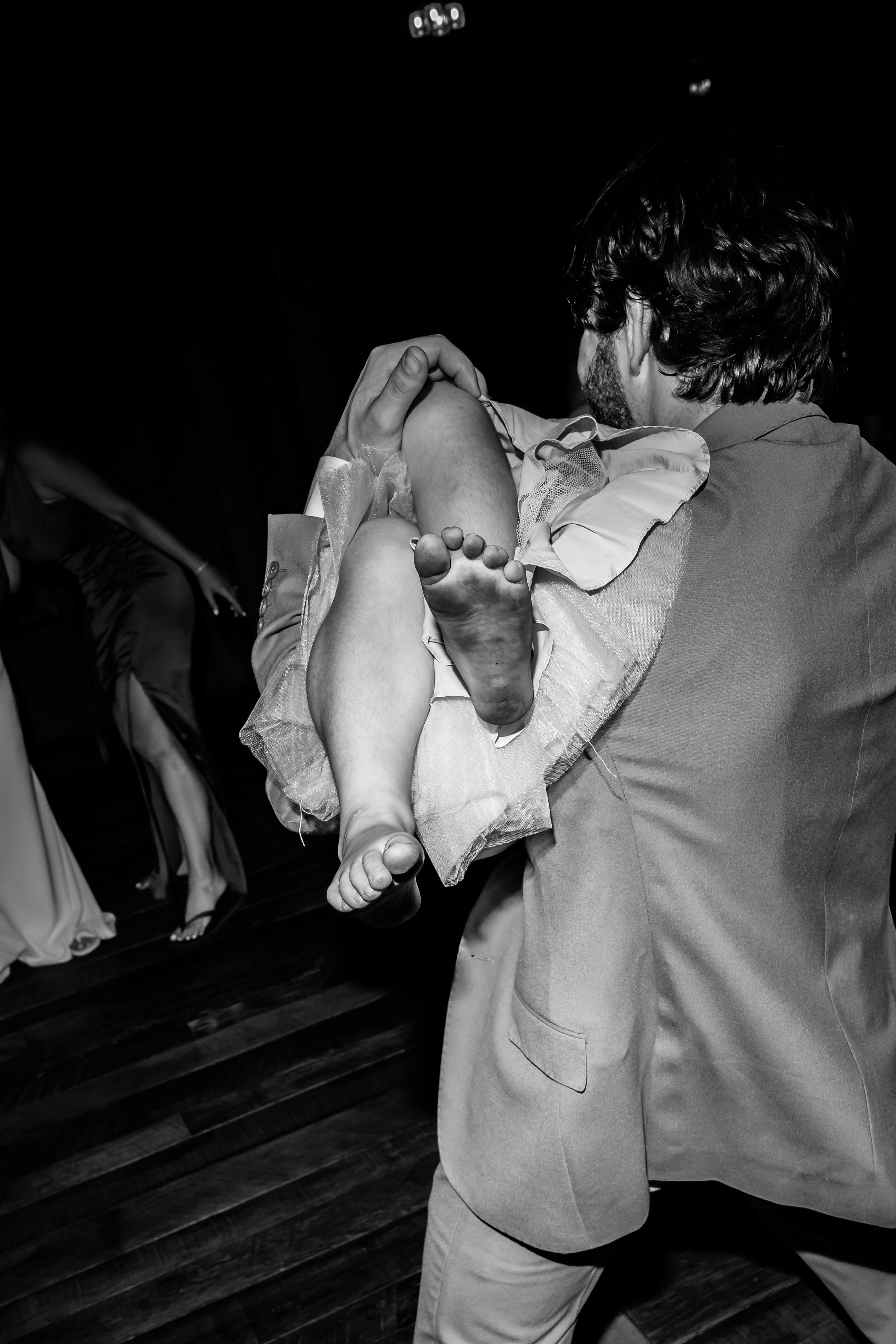 Black and white photo of groom carrying barefoot bride on wedding reception dance floor.
