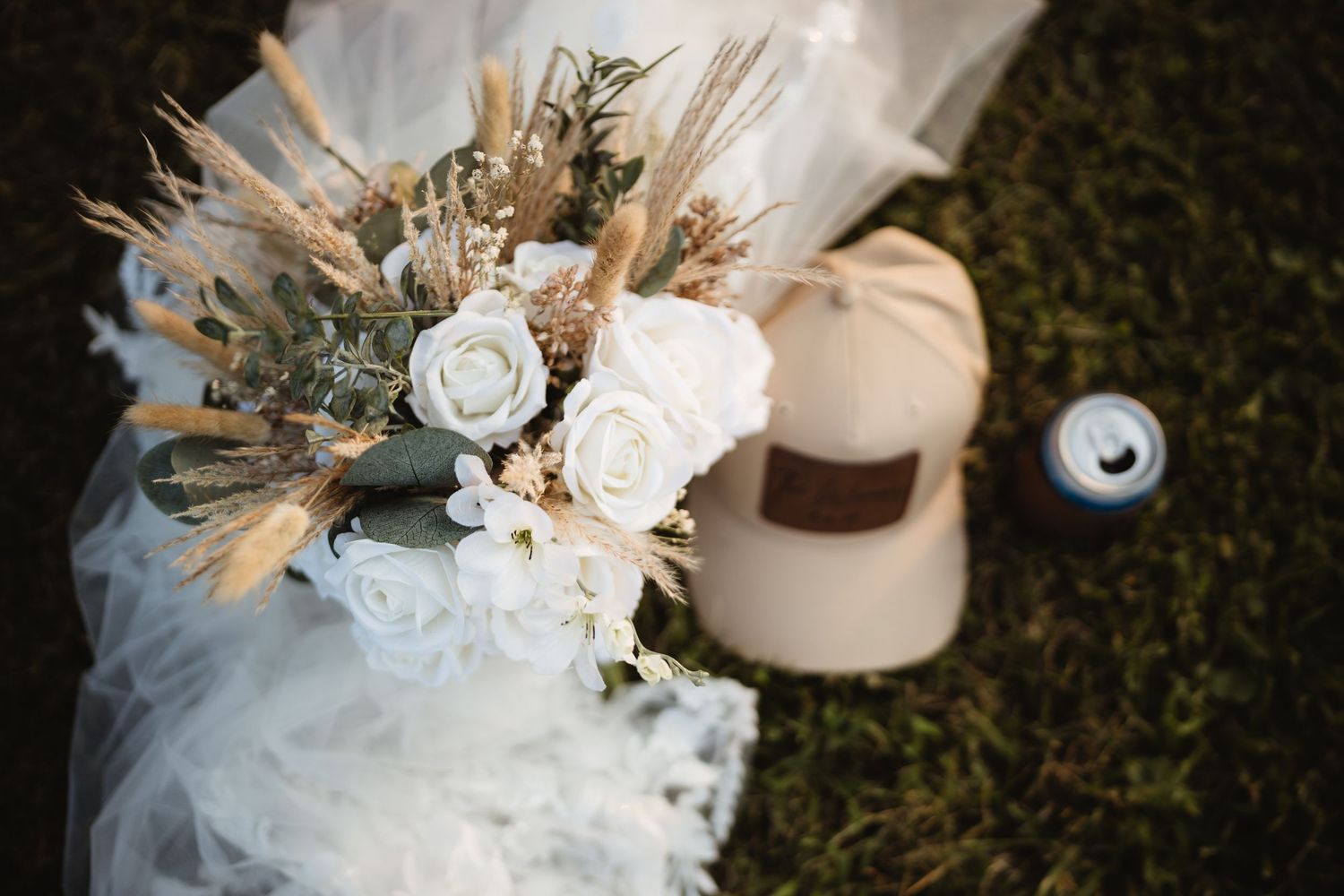 Rustic wedding bouquet with white roses and dried pampas grass beside a beige cap on green grass.