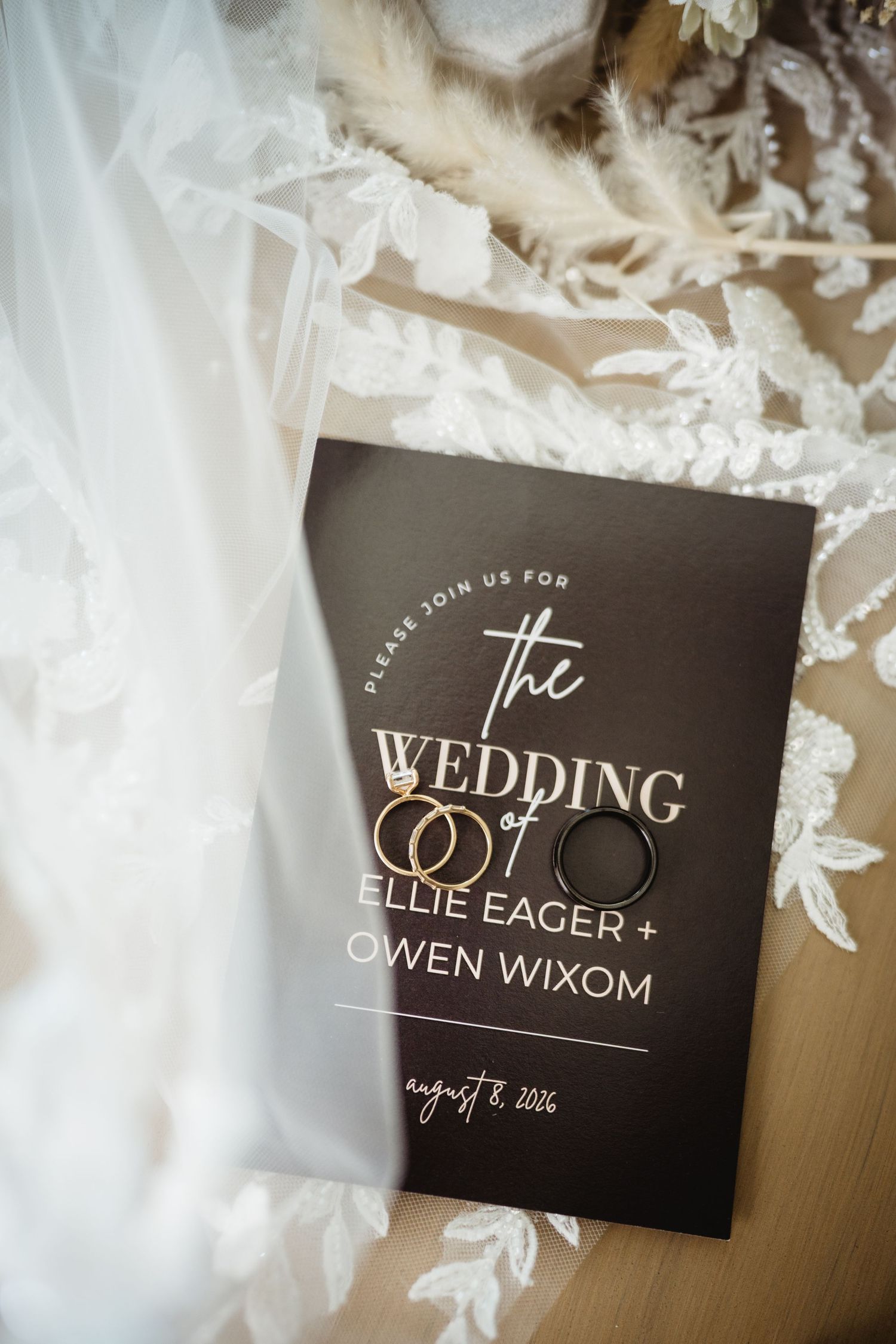 Wedding invitation for Ellie Eager and Owen Wixom on August 8, 2026, with gold rings resting on dark card beside lace bridal gown.