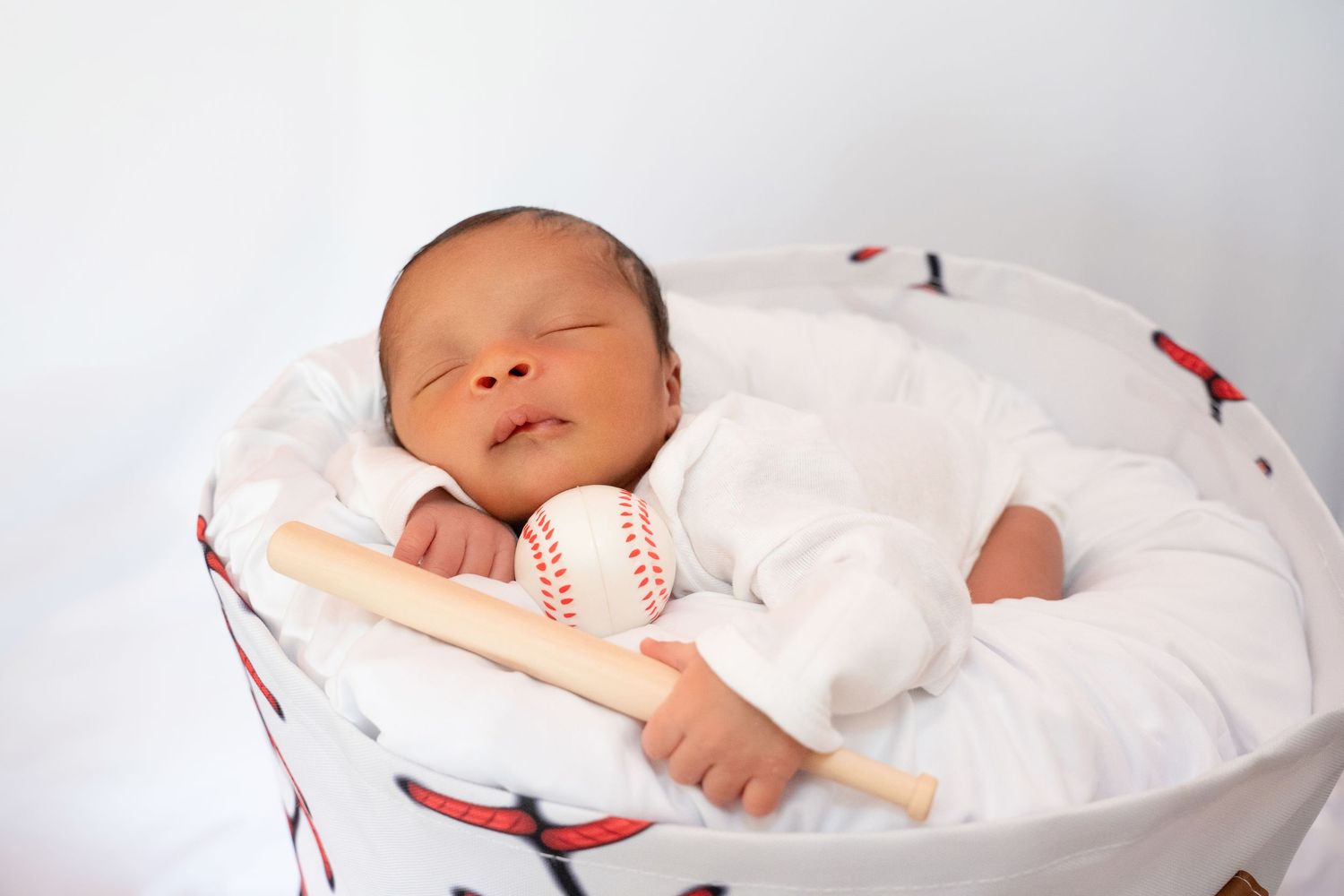 Peaceful newborn baby sleeping in baseball-themed bean bag holding a mini bat and baseball.