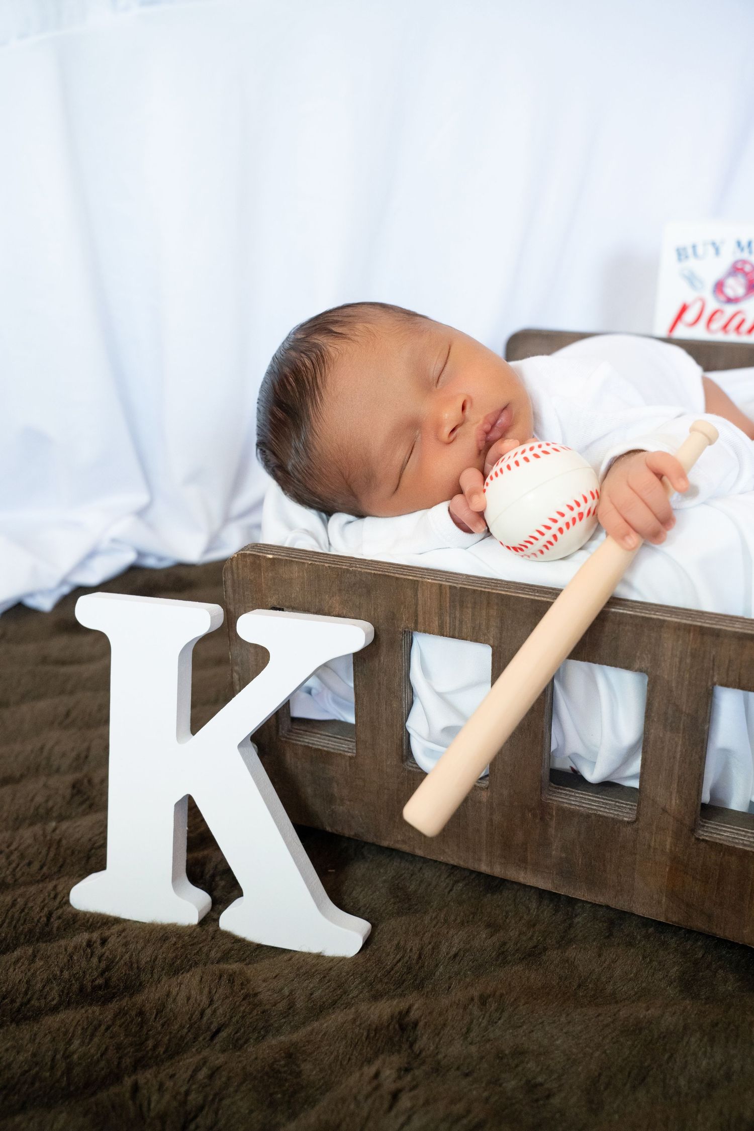 Sleeping newborn in wooden crib holding mini baseball bat with white letter K beside him.