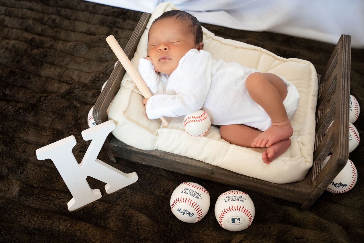 Newborn baby sleeping in wooden crate surrounded by baseballs and mini bat with letter K.