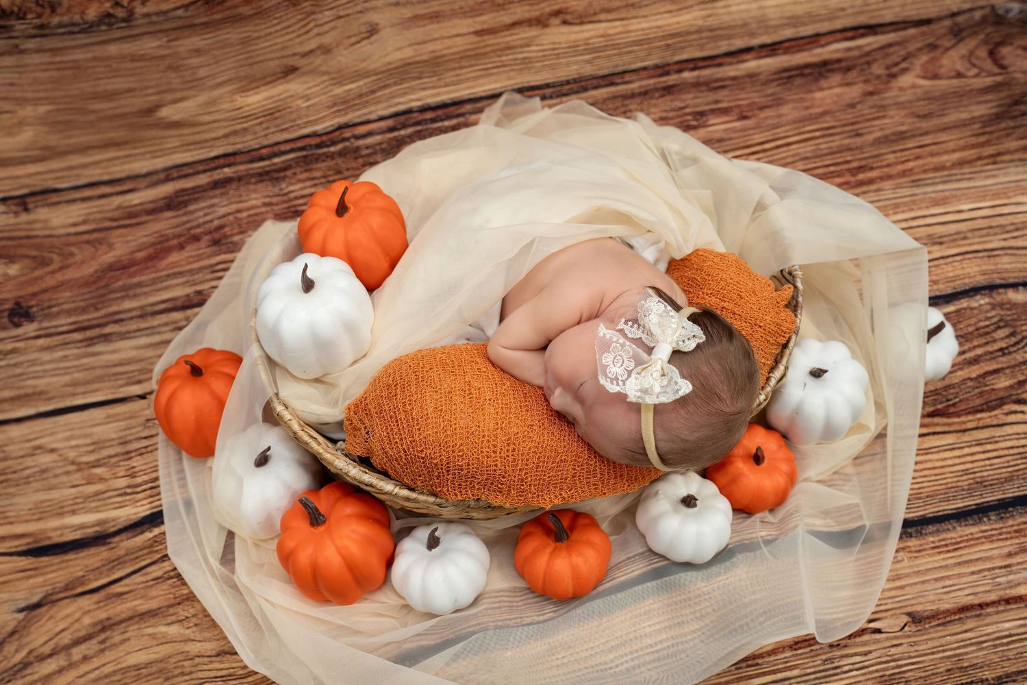 Sleeping newborn baby in a basket surrounded by orange and white pumpkins on wood.