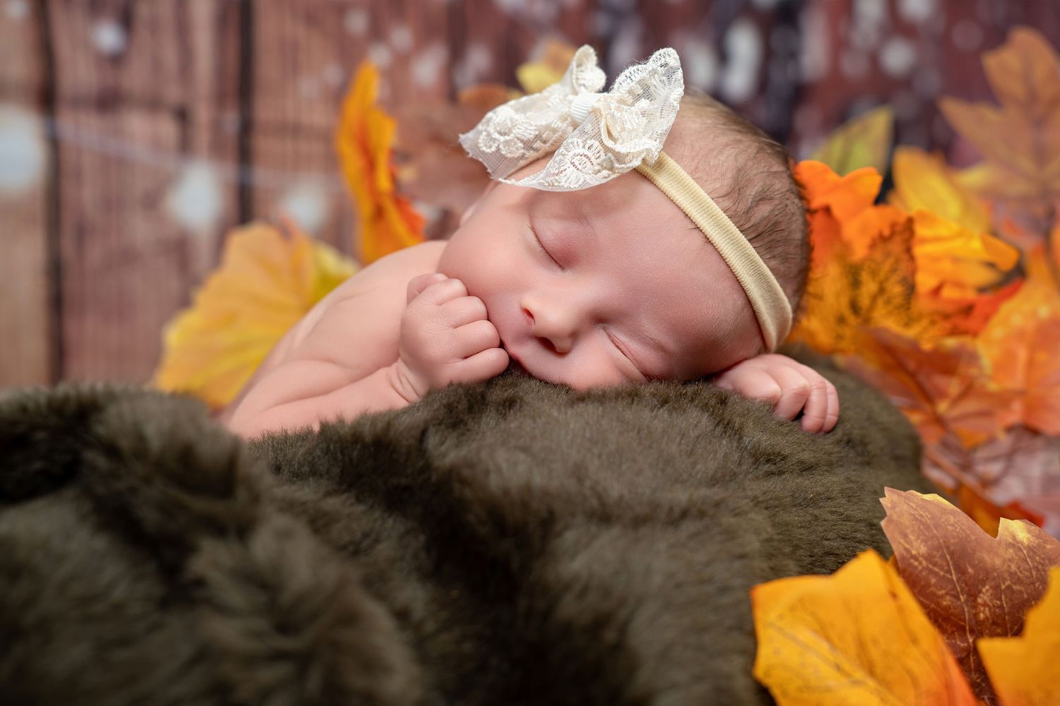 Newborn baby sleeping on brown fur blanket with autumn leaves and white lace headband.