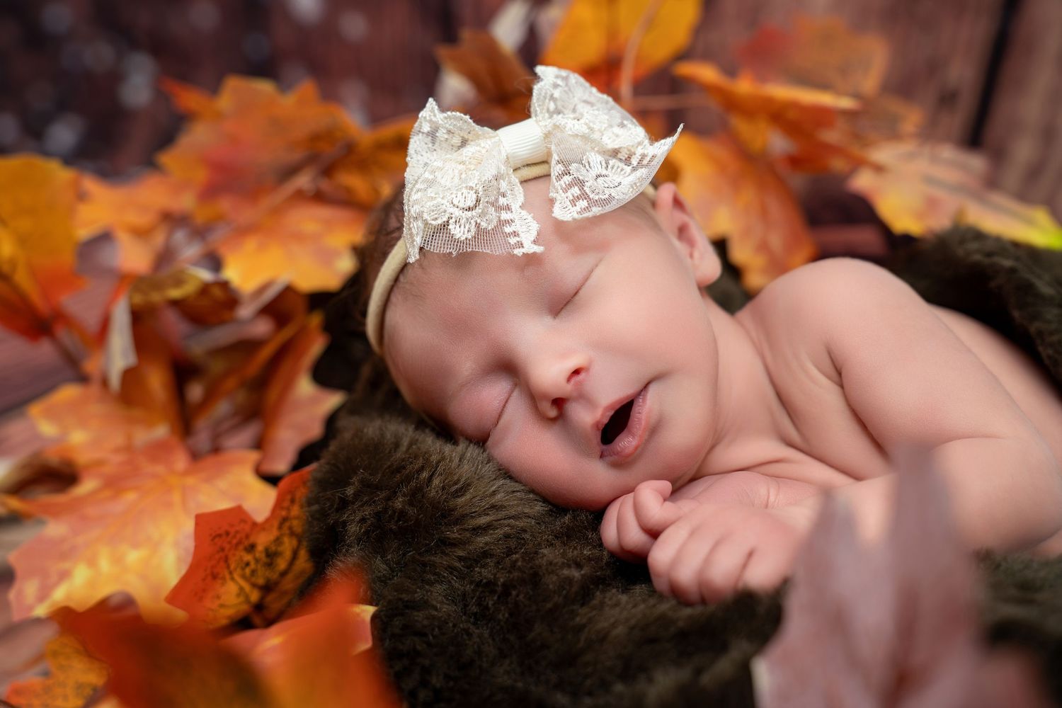 Close-up of sleeping newborn baby with lace bow headband among colorful fall leaves.