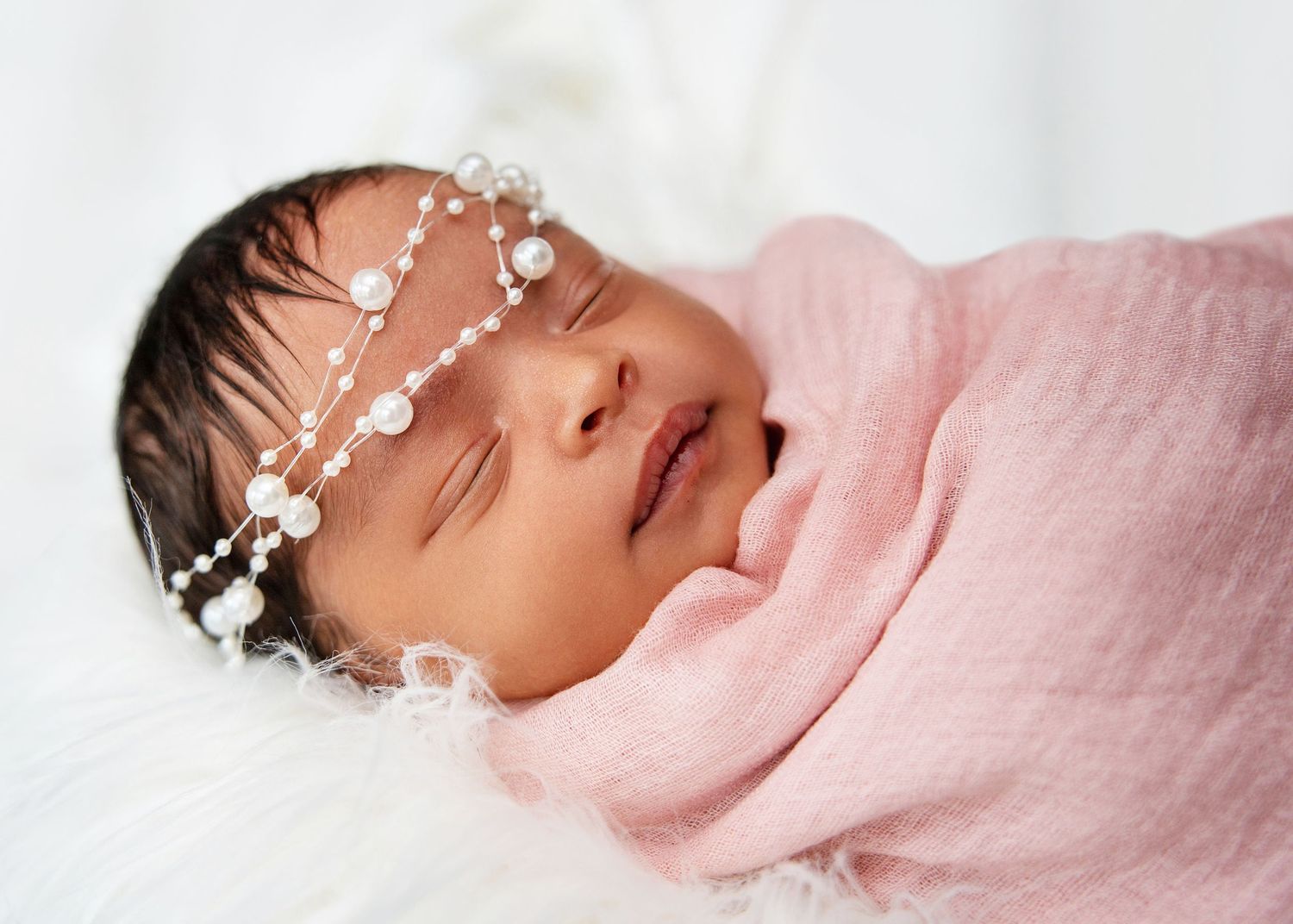Newborn baby swaddled in pink wrap with pearl headband sleeping peacefully.