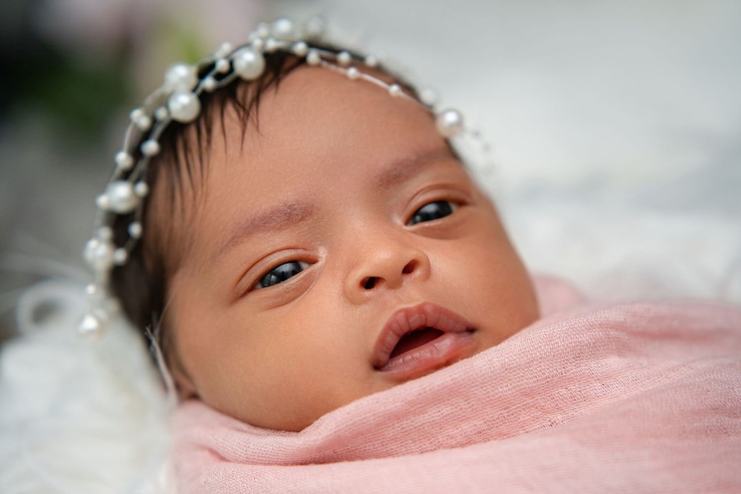 Awake newborn baby with pearl headband swaddled in pink looking at camera.