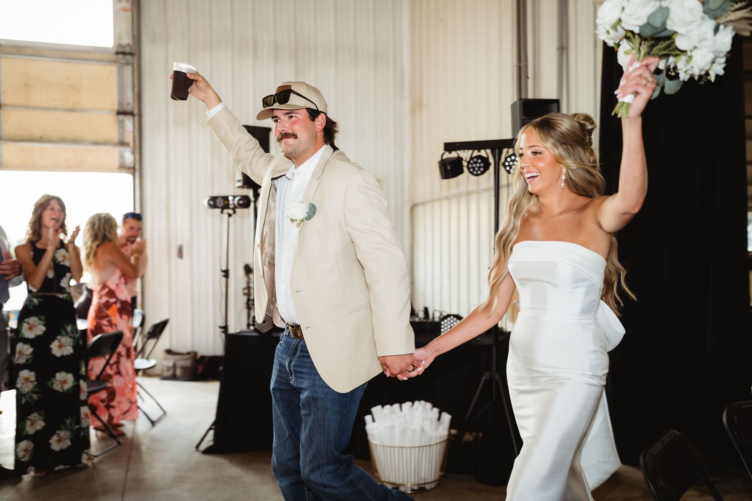 Bride in white jumpsuit and groom in tan blazer and jeans celebrate their wedding reception entrance.