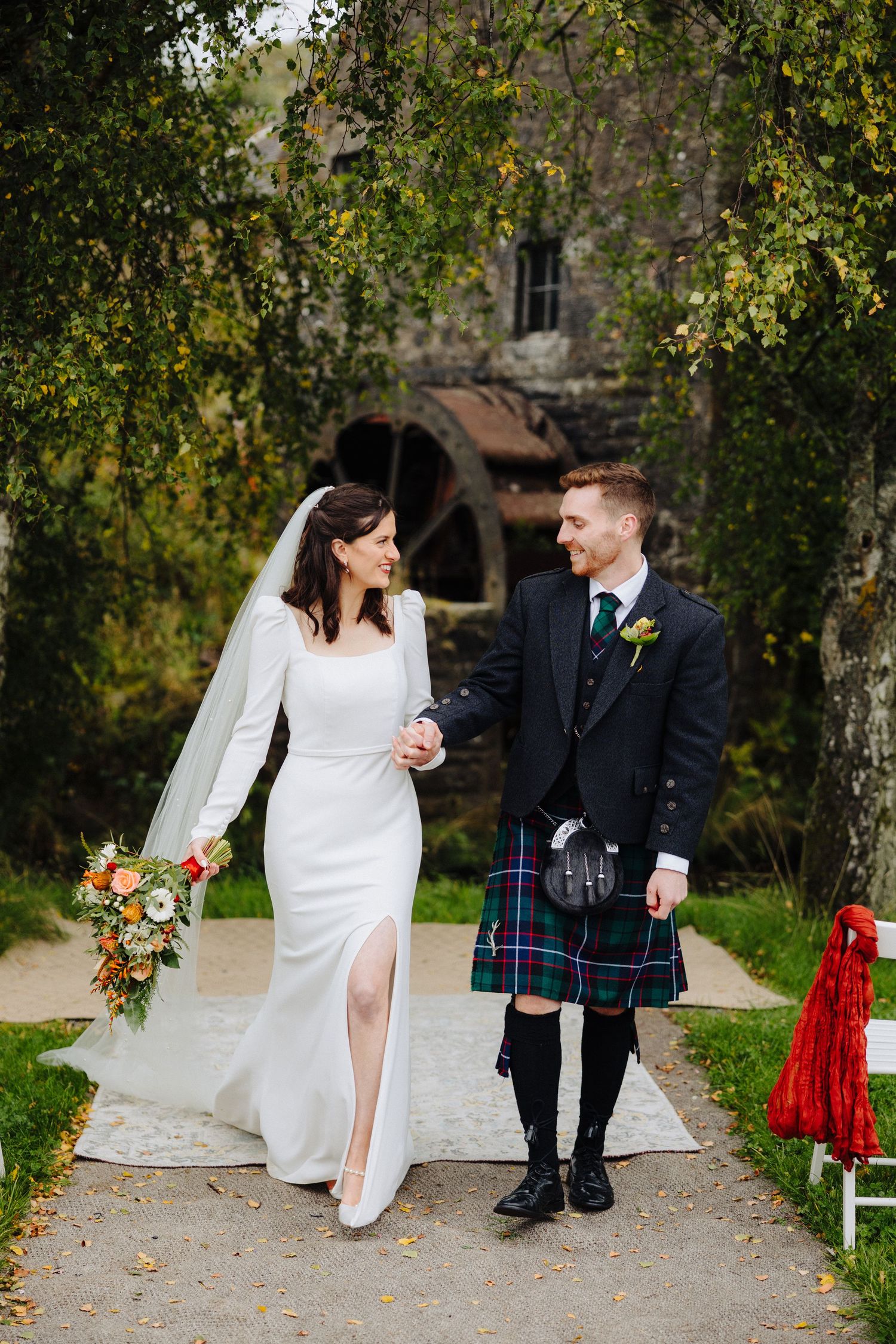 Bride holding autumn bouquet walks with kilt-wearing groom near rustic stone building in fall.