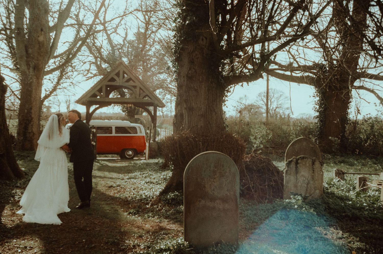 Bride and groom stand near a lychgate in a churchyard with a vintage red VW camper van in the background.