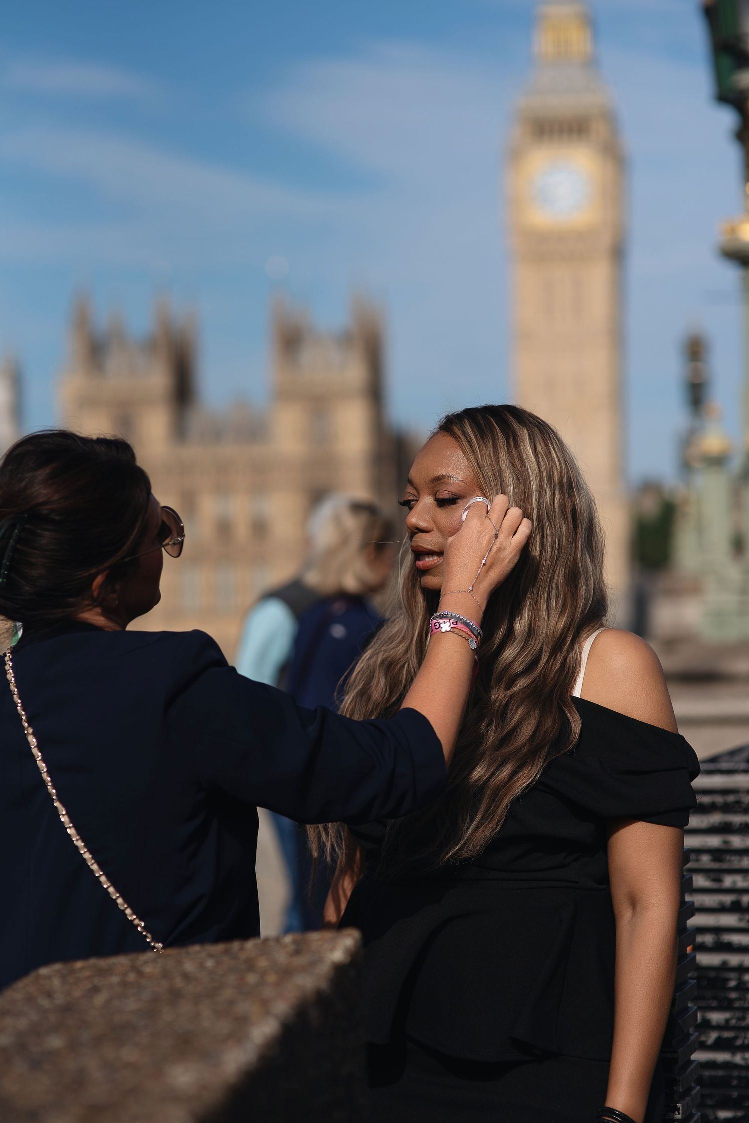 Two women near Big Ben in London, one touching the other's face, both wearing black outfits on a sunny day.