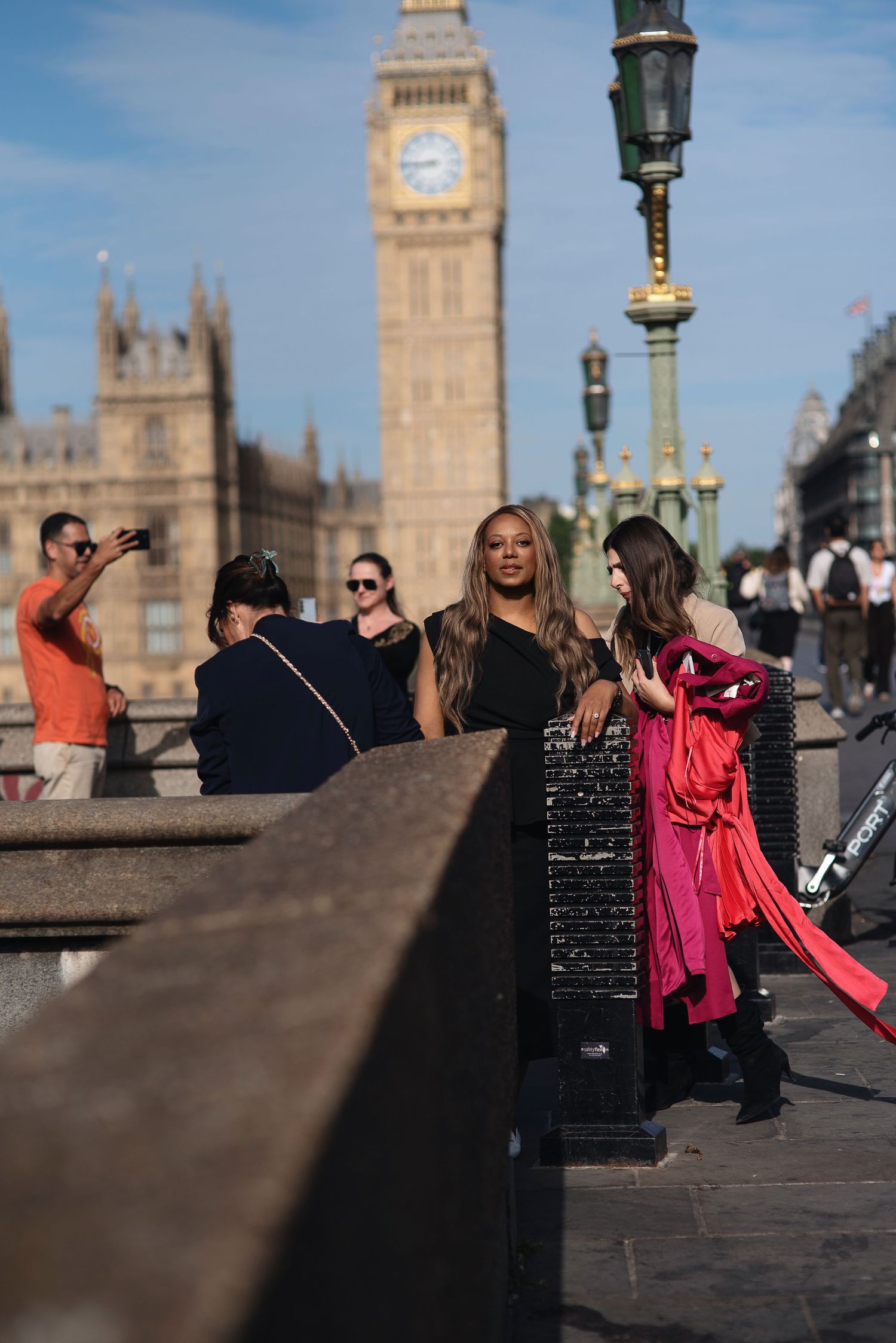 Two stylish women pose near Big Ben in London, one in black dress and another in pink outfit, on a sunny day.