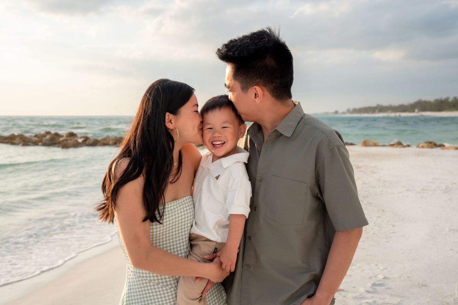 Loving Asian family at beach, parents kissing smiling toddler boy with ocean waves behind them.
