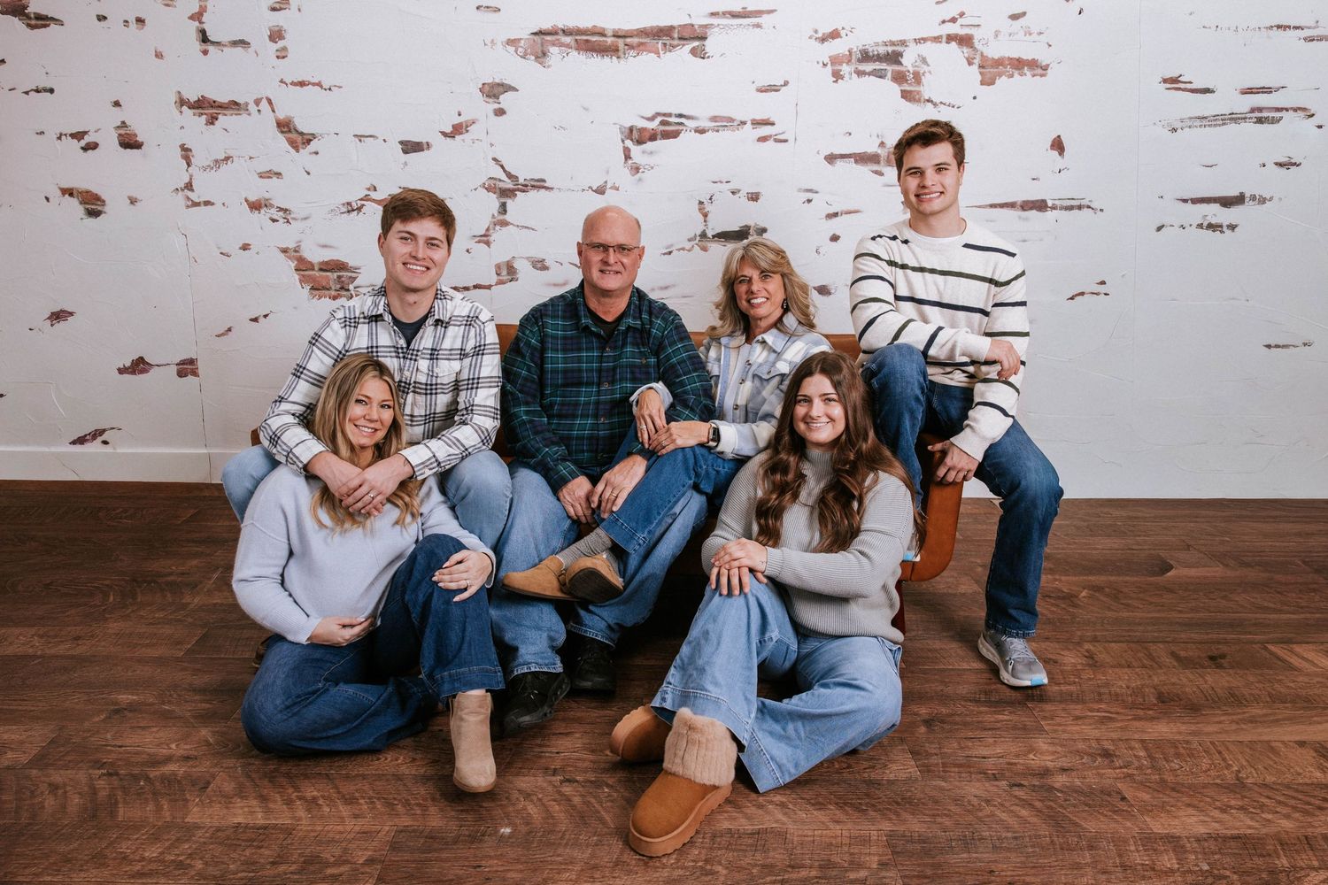 Family of six posing for a portrait, smiling warmly in casual attire against a rustic white brick wall.