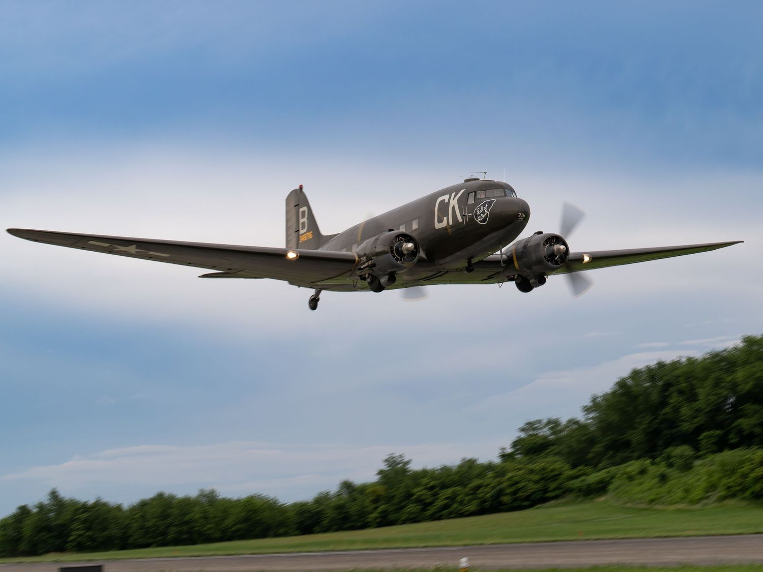Vintage WWII-era DC-3 military aircraft taking off against a cloudy sky, with green trees in the background.