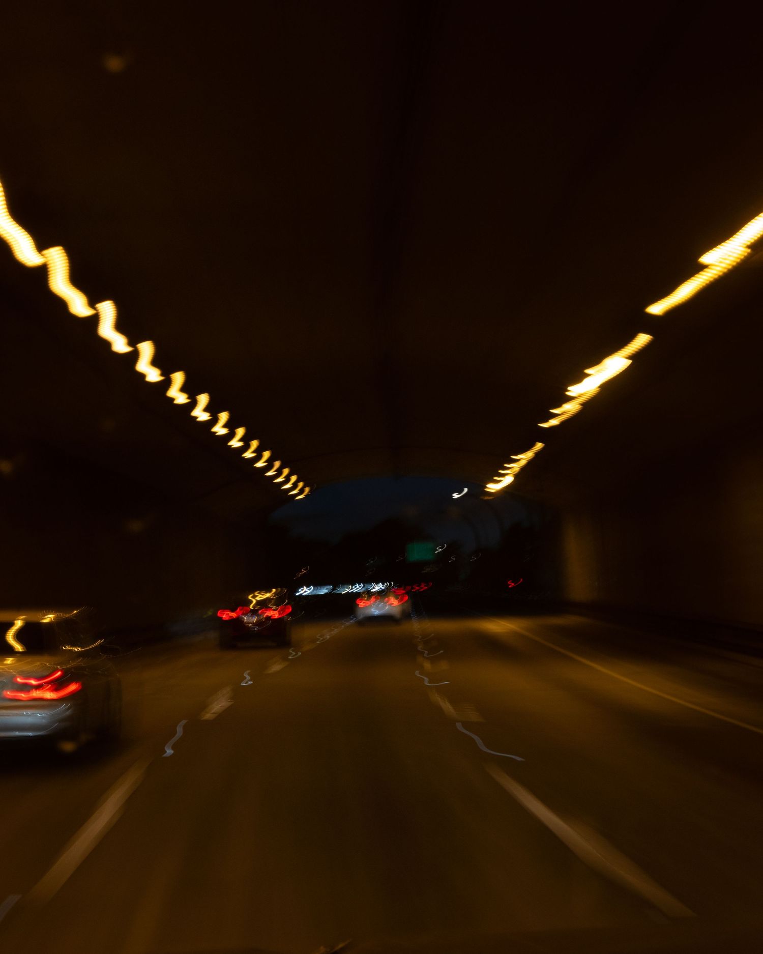 Blurry nighttime highway tunnel with glowing yellow lights lining the ceiling and red taillights of cars ahead.