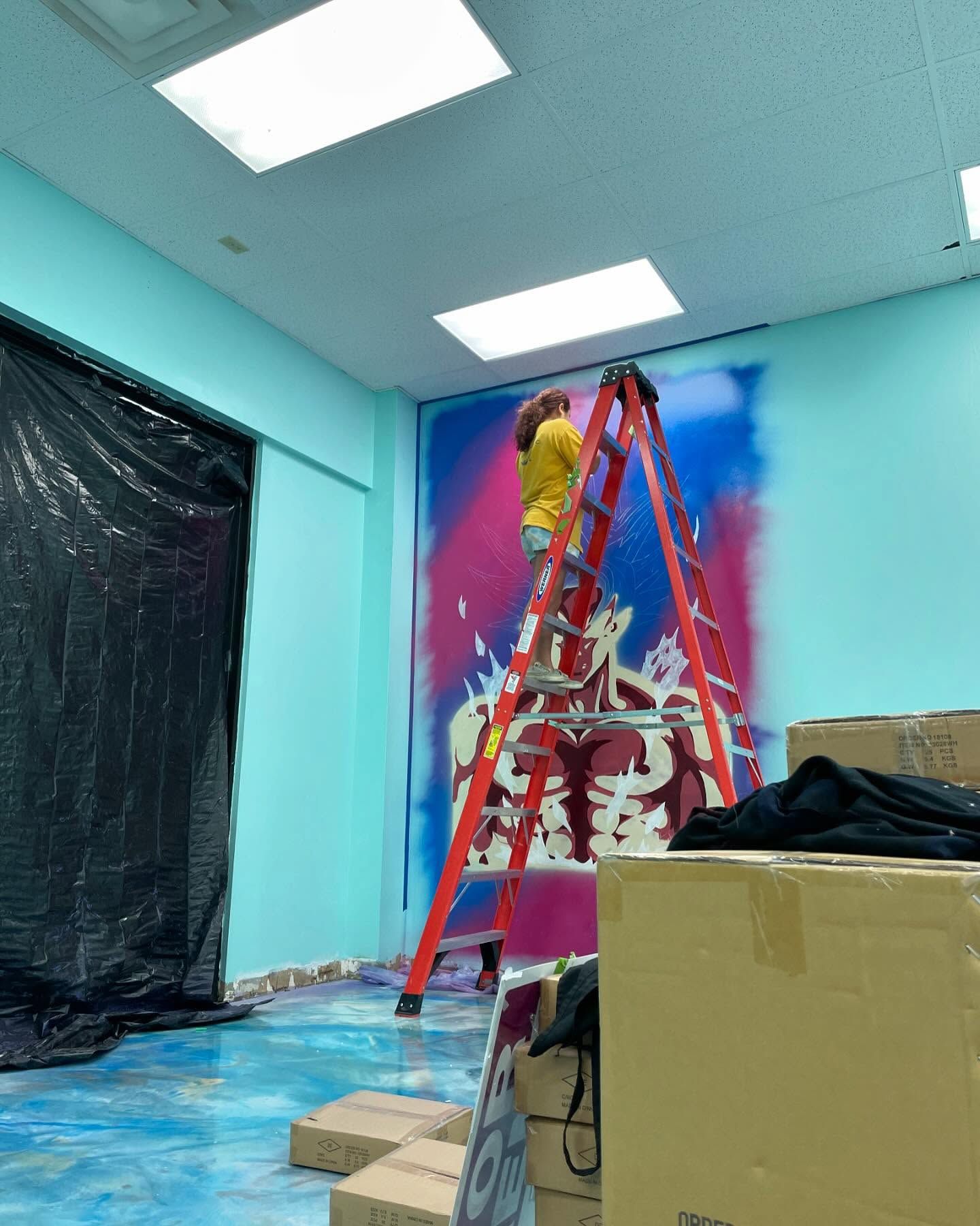 Artist on red ladder painting vibrant colorful mural on teal wall in commercial space under renovation.