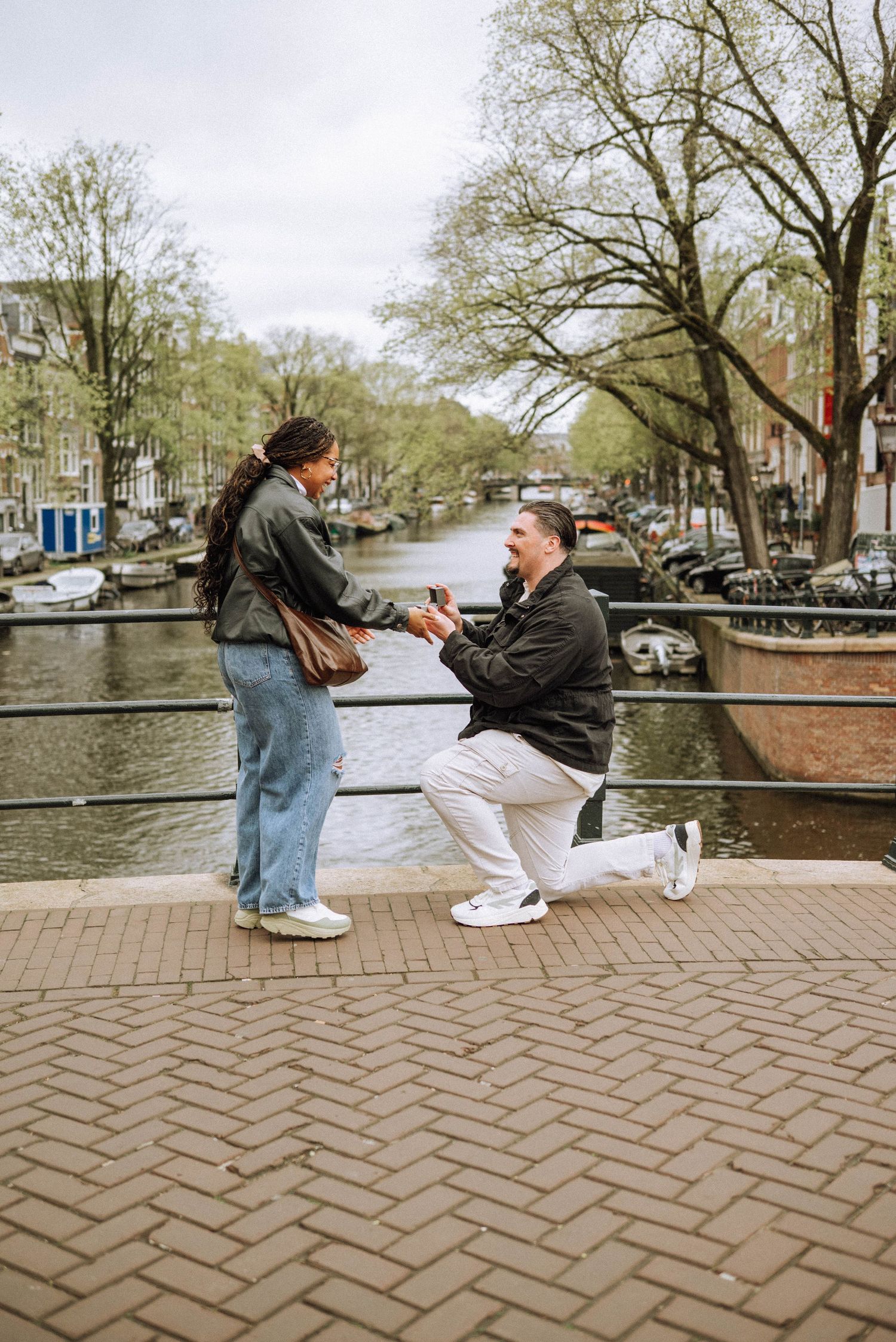 Man proposes on one knee to woman on a canal bridge in Amsterdam, with tree-lined waterway in background.
