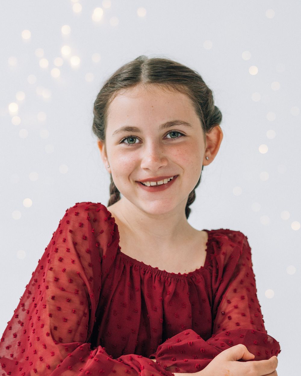 Smiling young girl with braids wearing a red long-sleeve dress, posing against a white bokeh background.