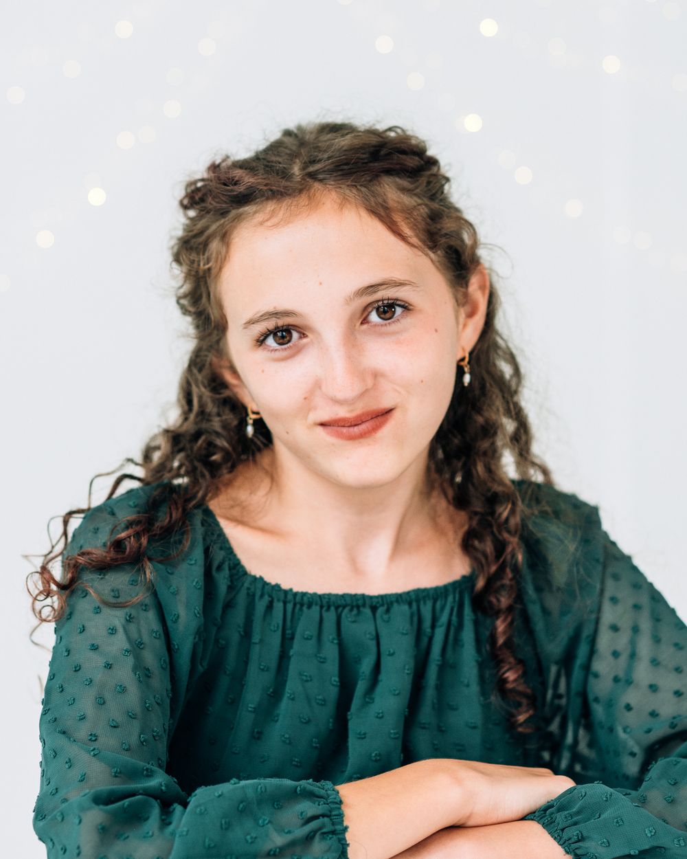Young woman with curly hair wearing a green textured blouse, smiling against a light bokeh background.