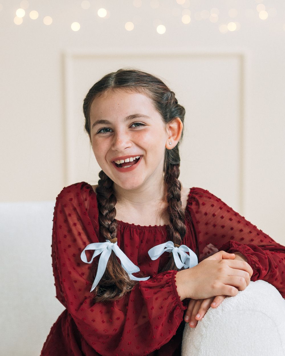 Smiling girl with braided pigtails tied with blue ribbons wearing a red dress, sitting against a white background.