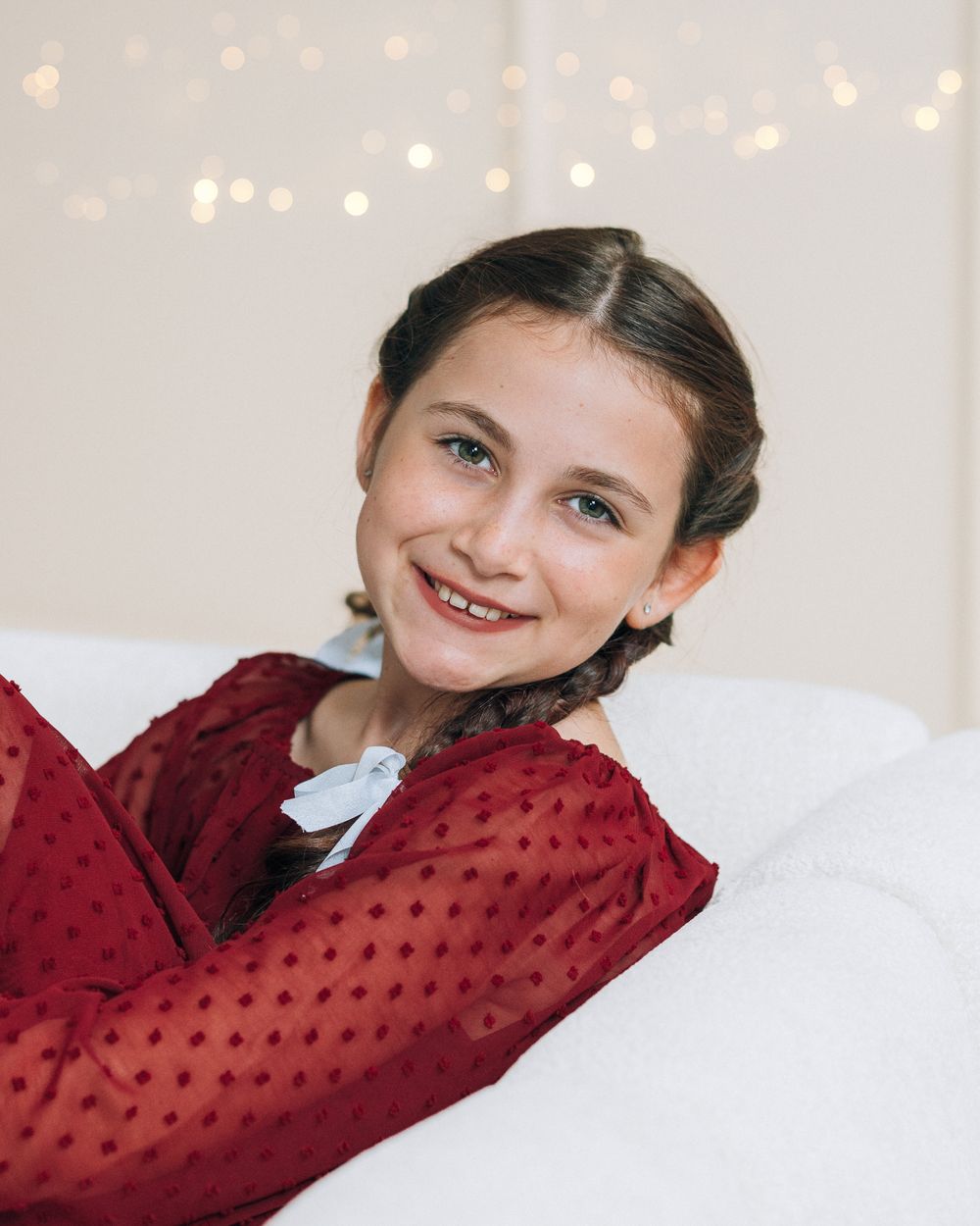 Smiling young girl in red polka dot dress leaning on white cushion with bokeh fairy lights background.