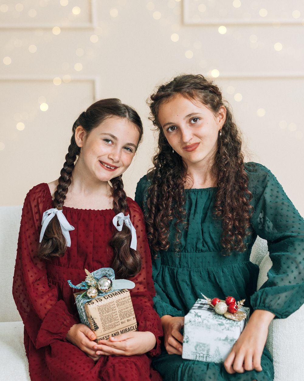 Two girls in red and green dresses smiling and holding Christmas gifts in front of bokeh lights.