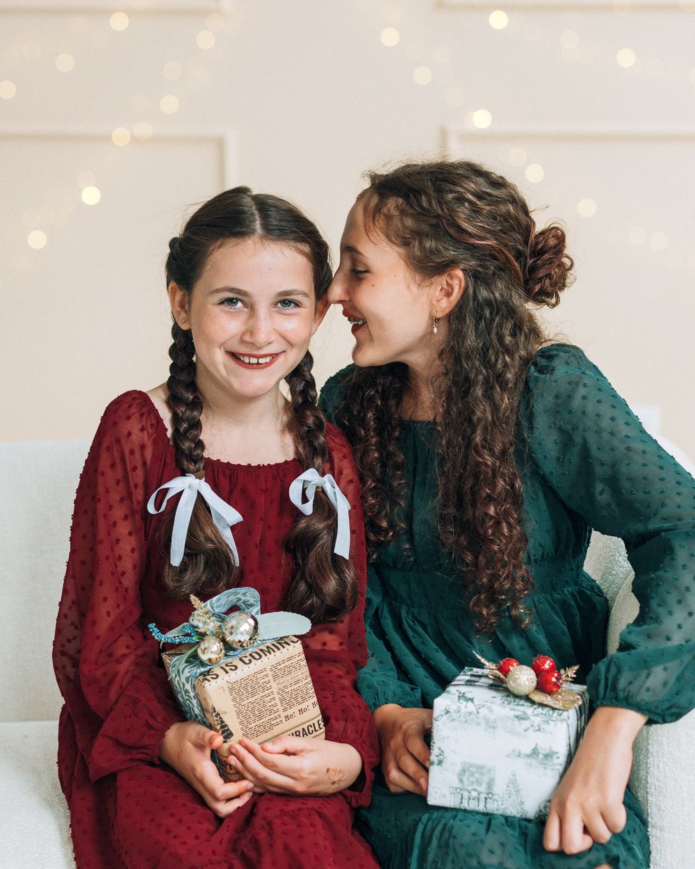 Two girls in festive red and green dresses exchange Christmas gifts while sitting together on a couch with bokeh lights.